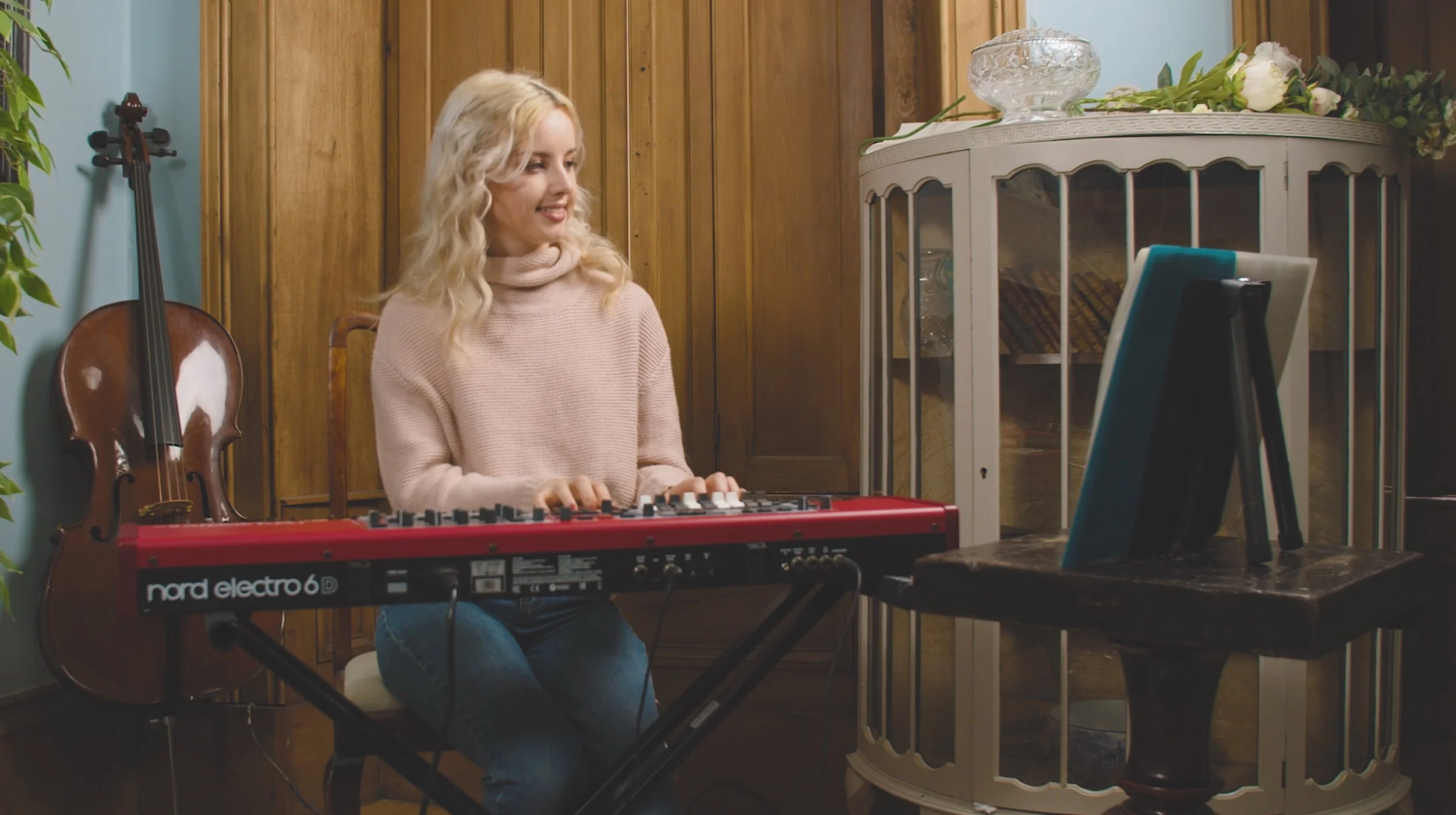 A woman with blonde hair playing a red Nord Electro 6D keyboard inside a room with wood paneled walls. Behind her is a cello leaning against the wall, and to her right is a small table with a framed photo and decorative items. The room has blue and wooden elements, with a white cabinet with glass doors in the background.
