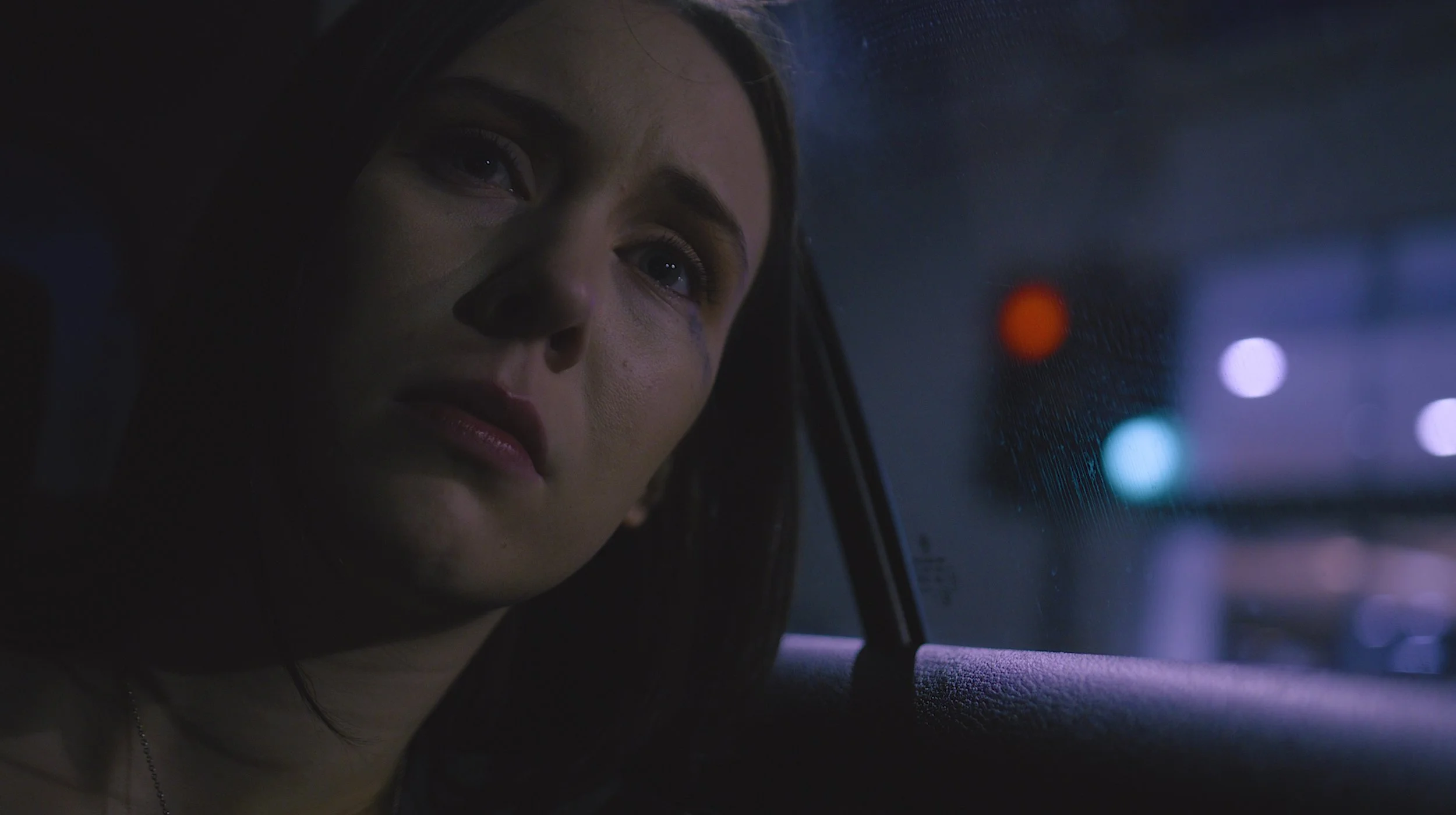 A woman with dark hair looking out of a car window at night, illuminated by city lights.