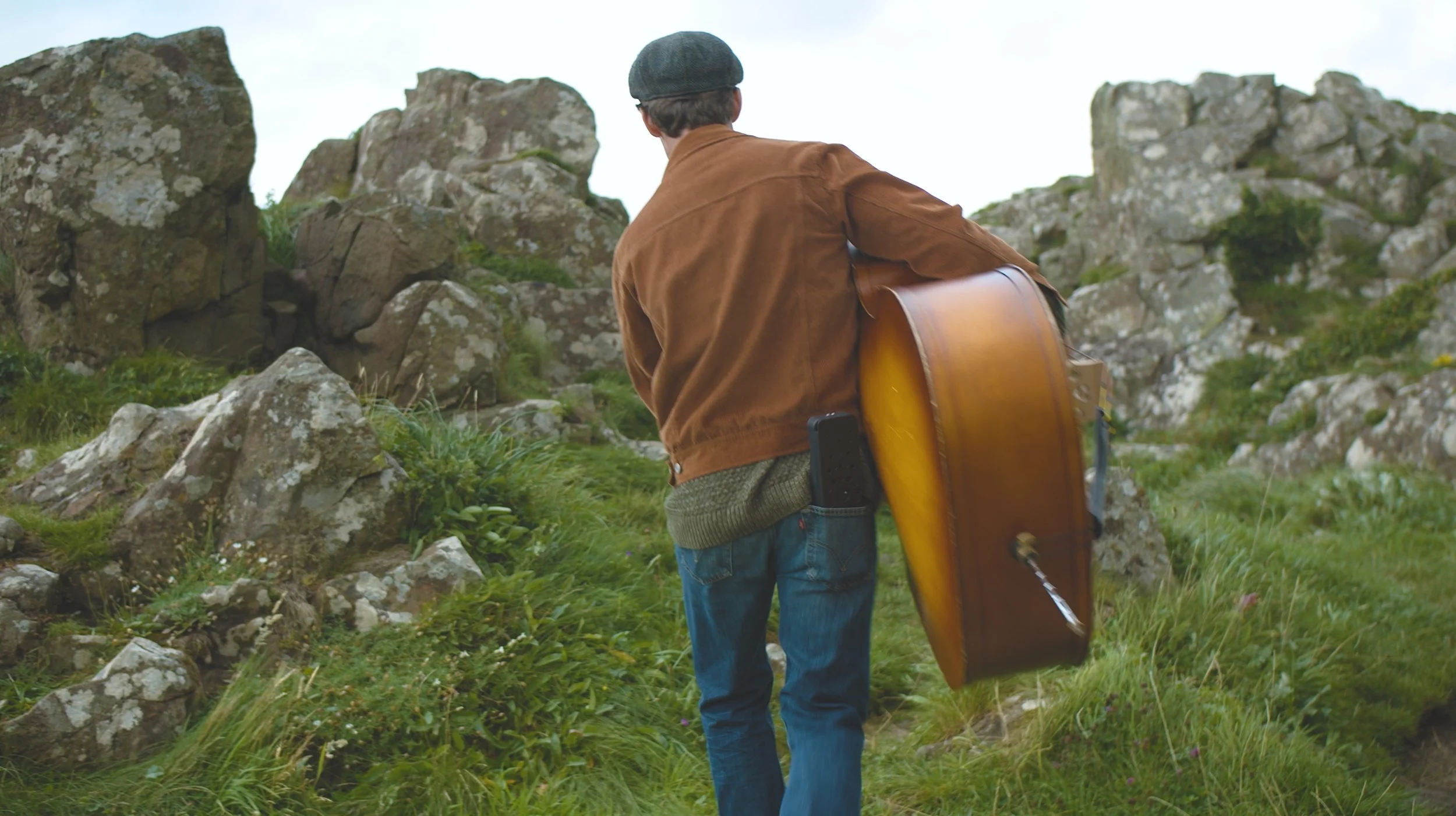 A person with a guitar in a green, rocky outdoor area, wearing a brown jacket, gray cap, and jeans, facing away from the camera.