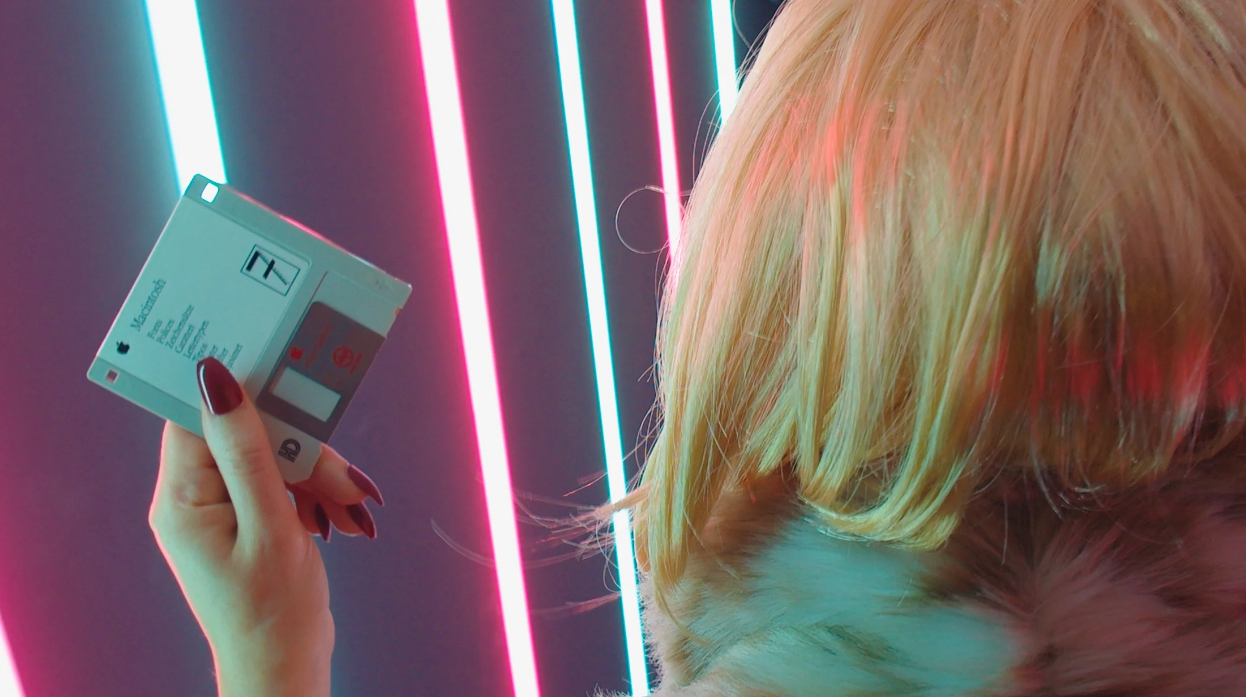 A person with long blonde hair holding a vintage Apple Macintosh 1 floppy disk. The background features neon pink and blue light stripes, giving a retro-futuristic aesthetic.
