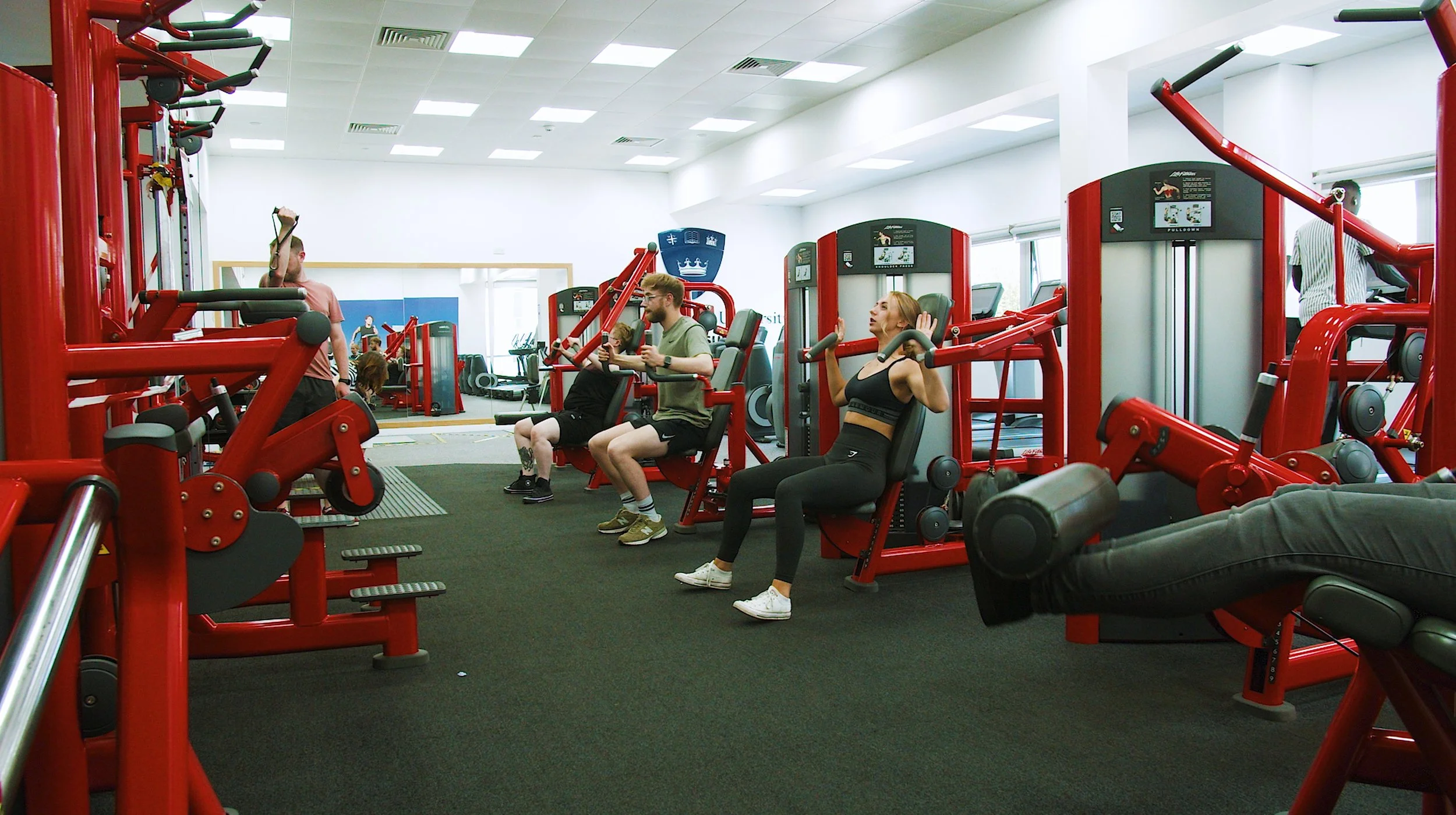 People working out on various exercise machines in a brightly lit gym with white walls and black flooring.