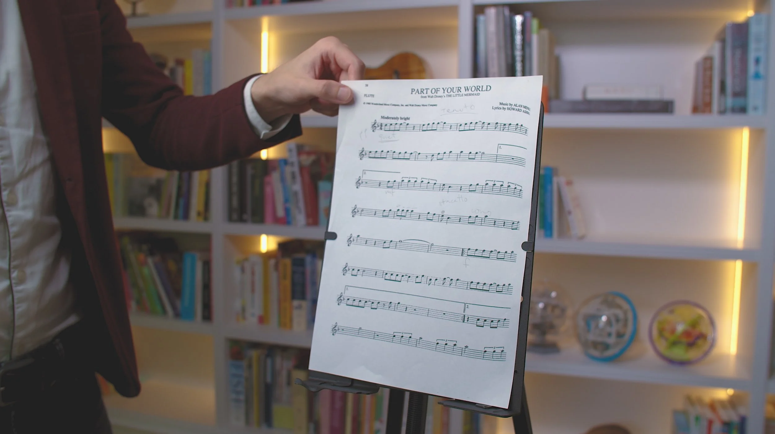 Person holding a sheet of music titled 'Part of Your World' with handwritten annotations, in front of a bookshelf.