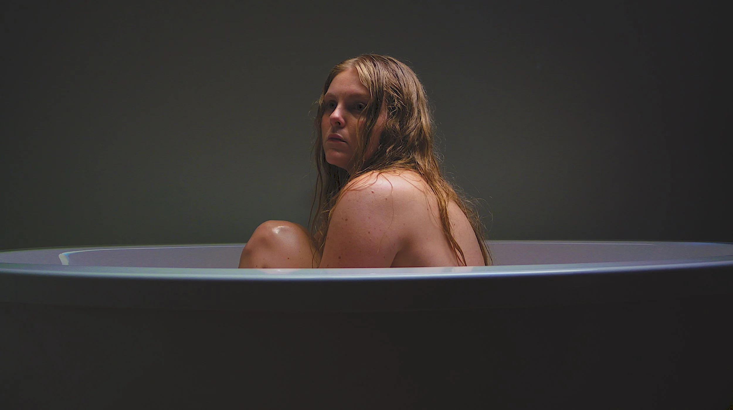 A woman with red hair sitting in a bathtub, looking over her shoulder with a neutral expression, in a dimly lit room.