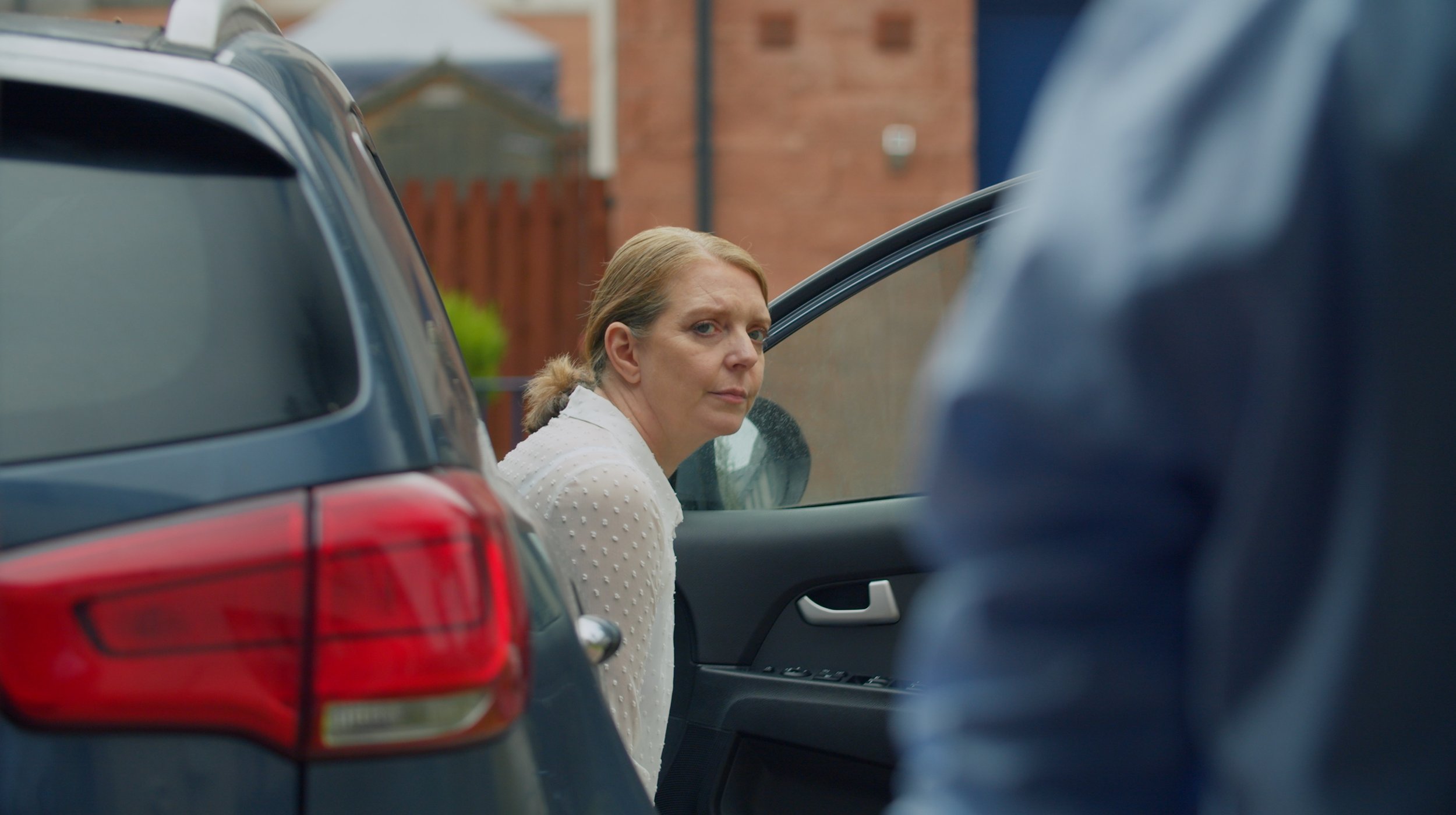 A woman with blonde hair wearing a white textured top, sitting in a car and looking towards the camera, seen through between parked cars on a suburban street with a brick house in the background.