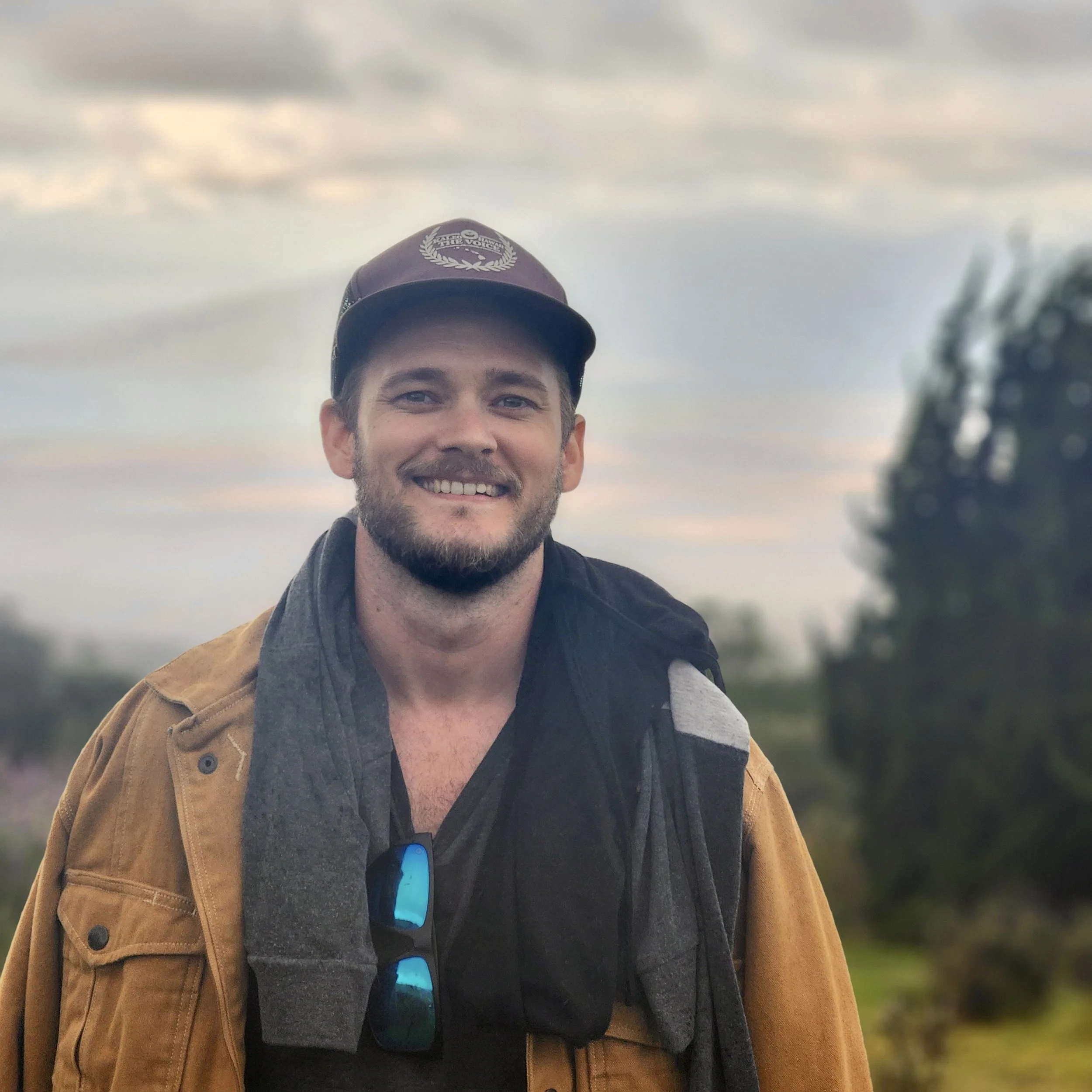 A man wearing a baseball cap and jacket, smiling outdoors with trees and sky in the background.