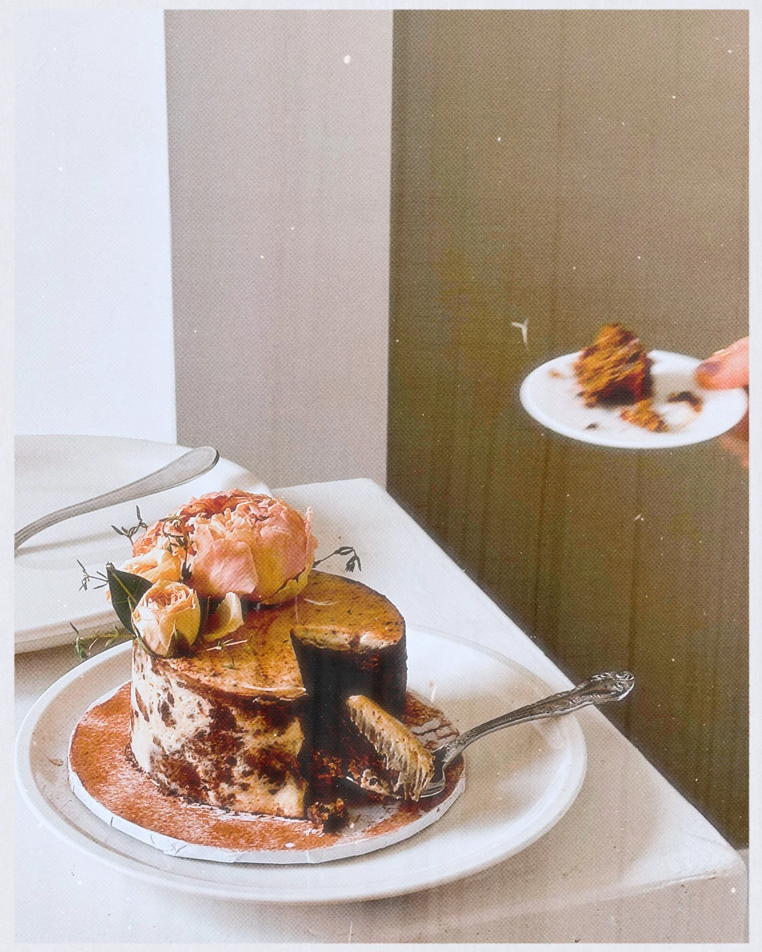 A caramel cake with a slice cut out, decorated with a pink flower and small berries, on a white plate on a table.