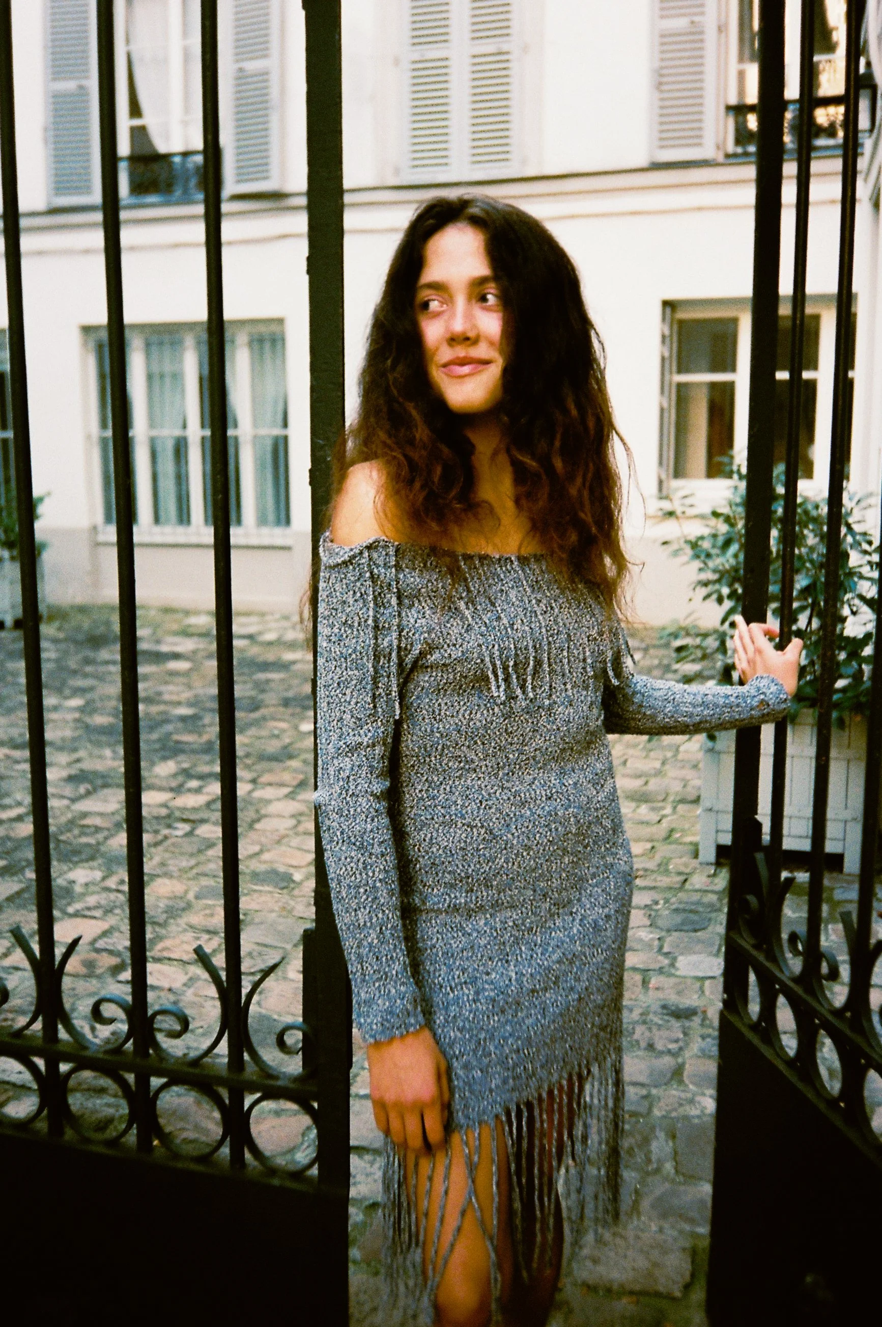 A young woman with long, wavy brown hair standing partially inside a black wrought iron gate on a cobblestone street, wearing a gray off-the-shoulder dress with fringe details at the hem, smiling and looking to the side while holding the gate.