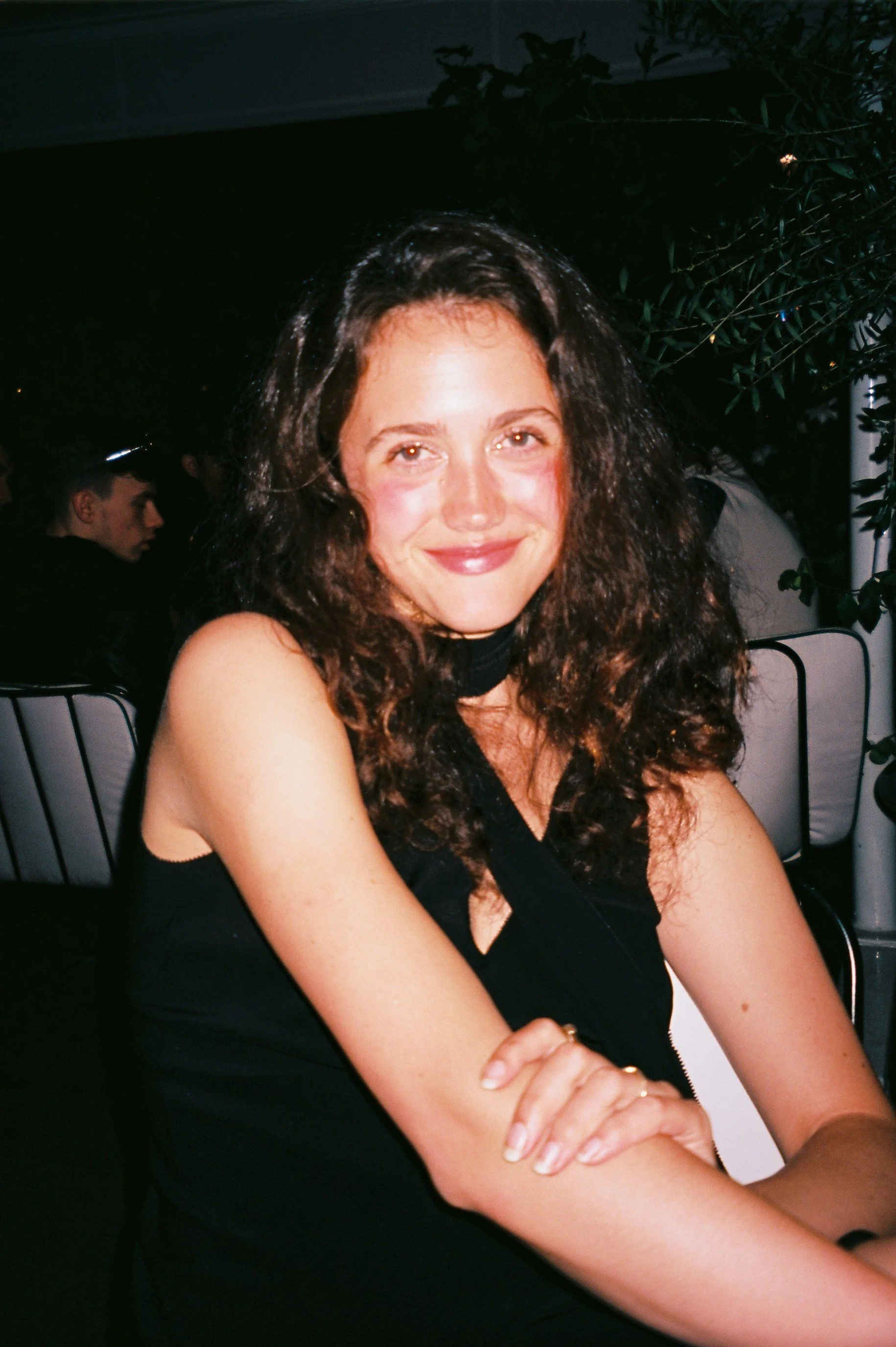 A woman with curly brown hair and light skin, smiling and sitting with her arms crossed, wearing a black sleeveless top.