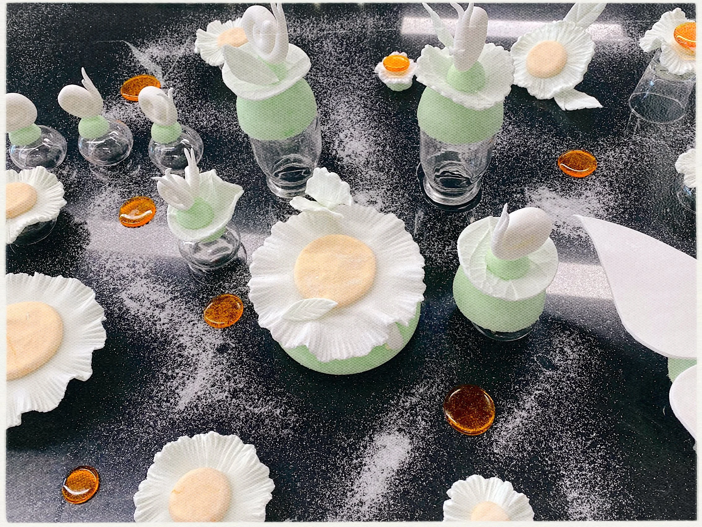 Decorative cookies shaped like daisies with white icing petals and orange centers, arranged on a black surface with scattered white powdered sugar and small amber-colored glass stones, accompanied by cupcake-like containers with pastel green bases and white icing, some topped with white fondant bunny ears and other decorative white flowers.