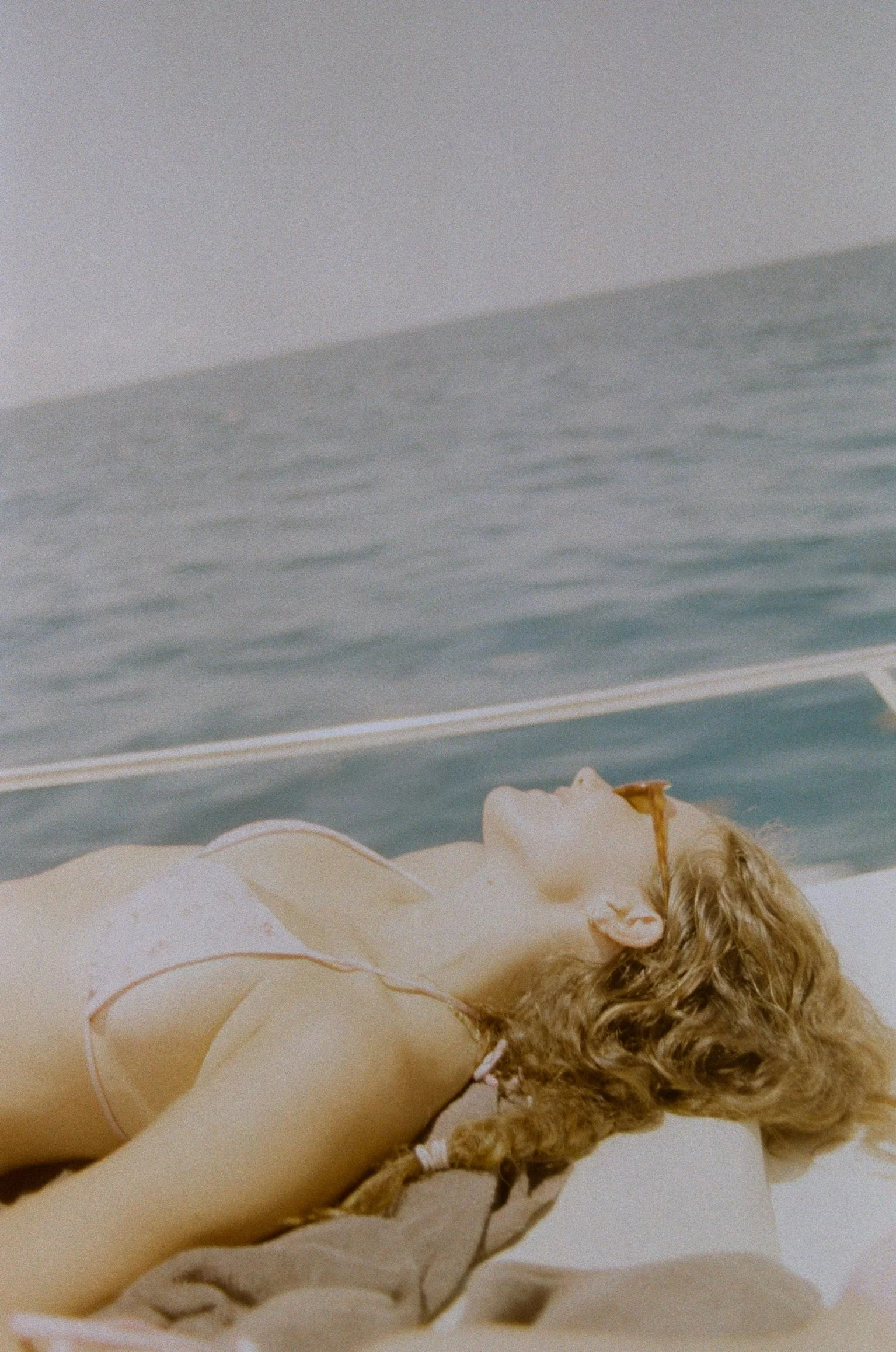 A woman with curly blonde hair wearing sunglasses sunbathing on a boat with the ocean in the background.