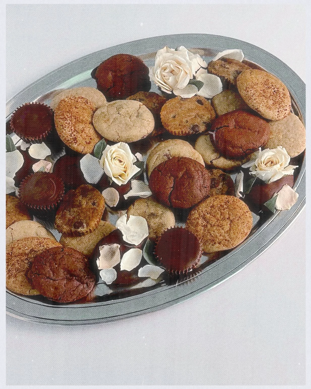 Assorted cookies and cupcakes on a silver platter with white rose decorations and flower petals.