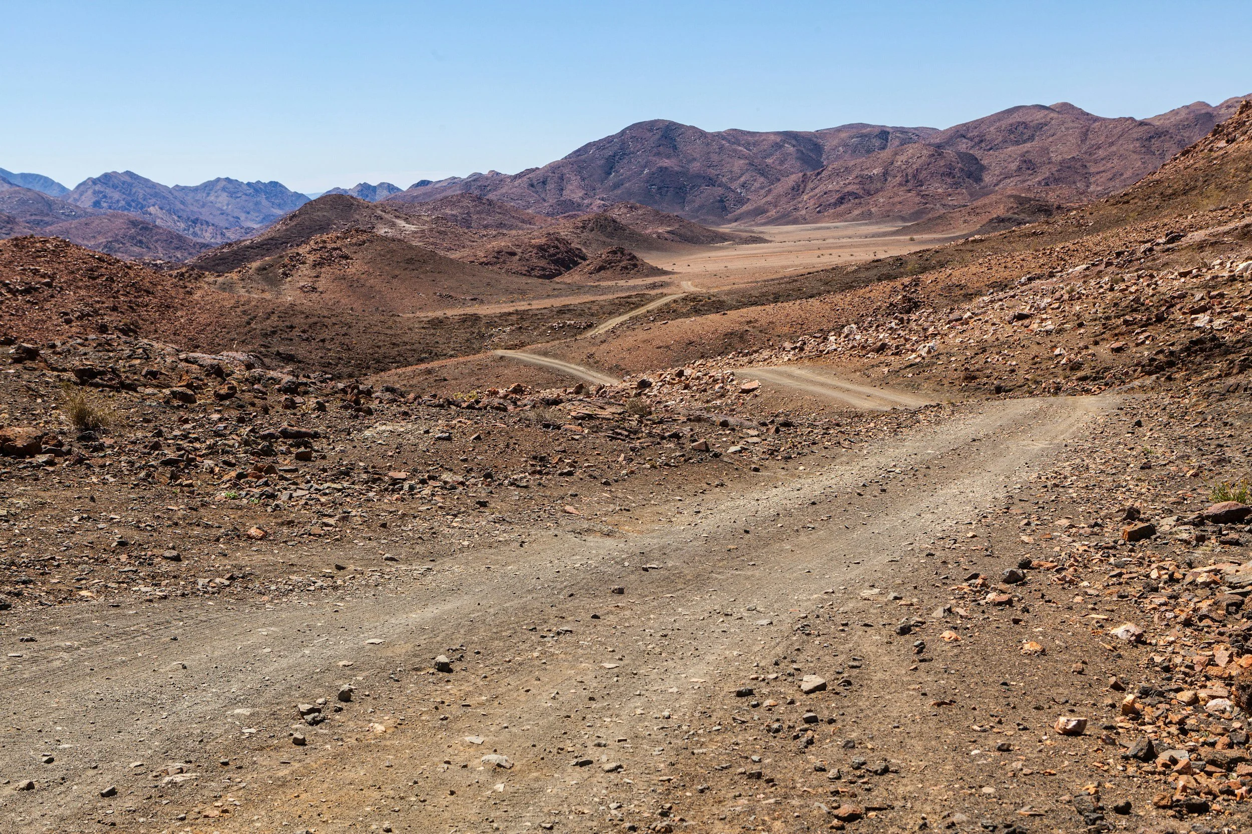 A dry, rocky desert landscape with a winding dirt road leading into distant hills under a clear blue sky.