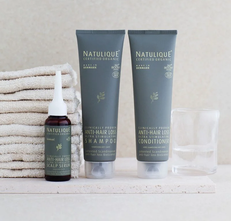 Natulique anti-hair loss products including shampoo, conditioner, and scalp serum, next to folded towels and an empty glass on a countertop.