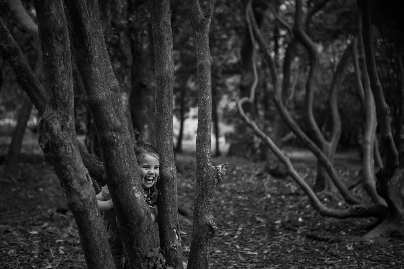 sister hides from her sibling and pokes head through trees at Cardiff Park whilst laughing
