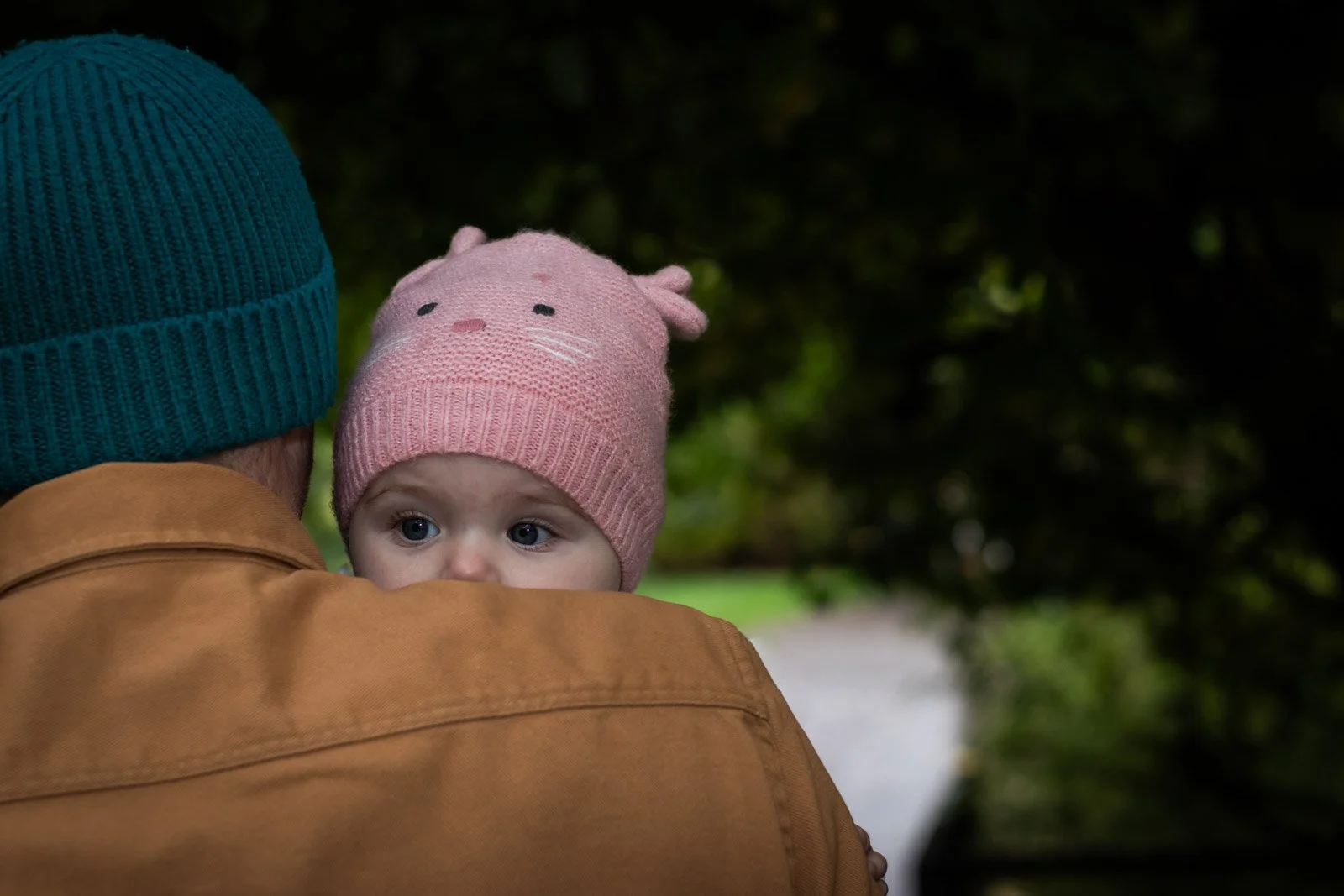 baby girl looks over fathers shoulder whilst held. Both wearing hats in the autumn weather