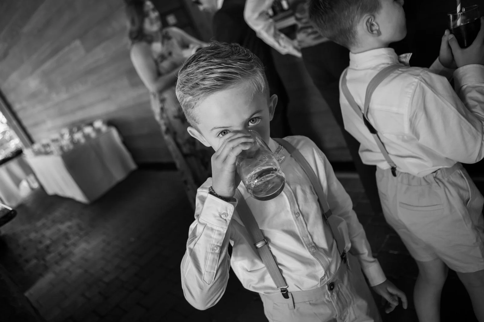 little boy at wedding looks over drinking glass as guests move around him