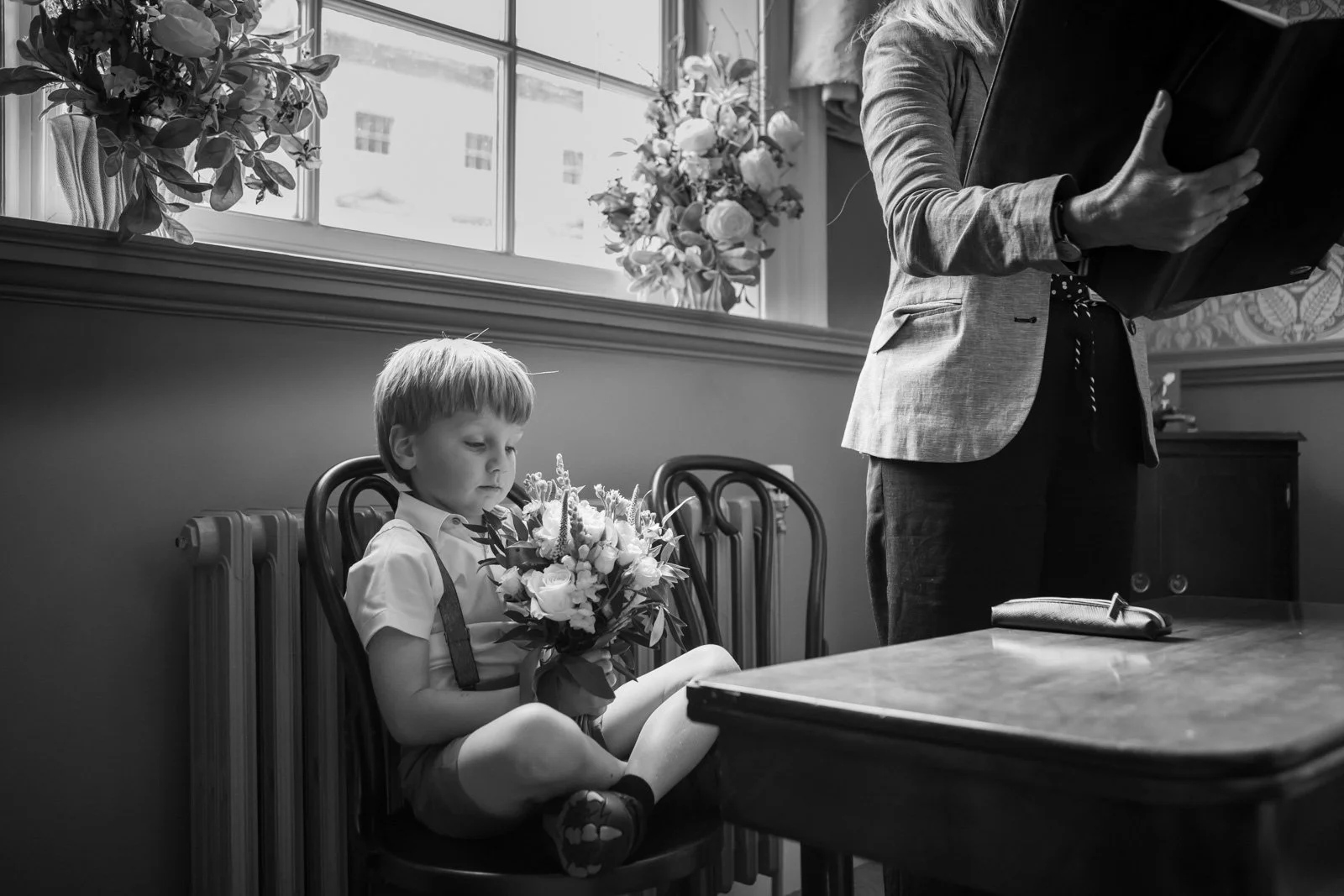 page boy holds a bouquet sat in chair at the front of ceremony as registrar conducts marriage