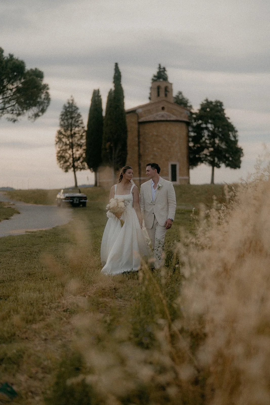 Wedding couple walking outside on grass, woman in a white dress holding a bouquet, man in a light-colored suit, church and trees in background, overcast sky.