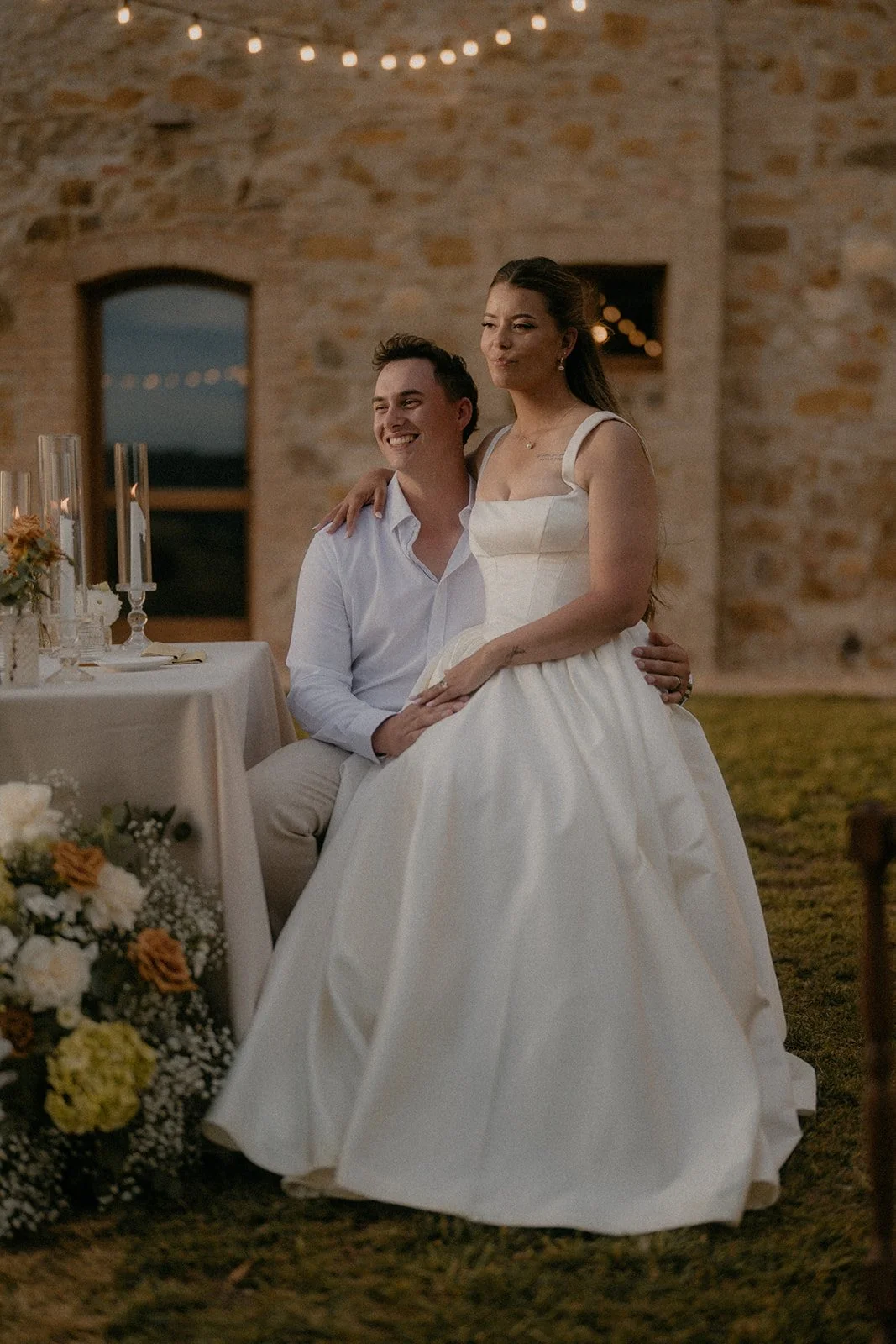 A newlywed couple at their wedding reception, with the groom sitting on a chair and the bride sitting on his lap, smiling and making a playful face. They are outdoors at dusk, with a rustic stone building and string lights in the background, and a de