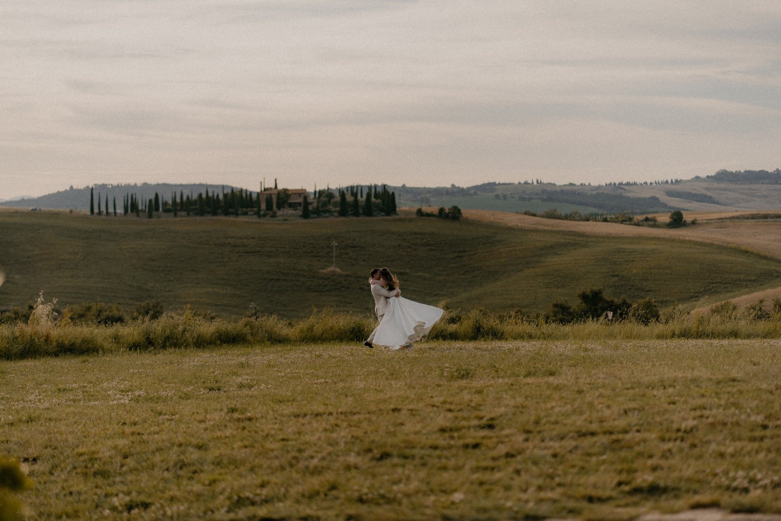 A couple dancing in a field with rolling hills and trees in the background, the woman in a white dress and the man in a light-colored suit, during a sunset or late afternoon.