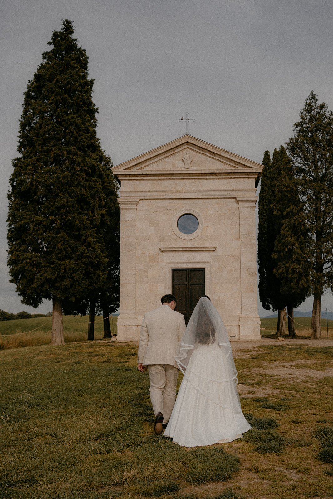 A newlywed couple walking away from the camera towards a small stone chapel, surrounded by trees, during sunset.