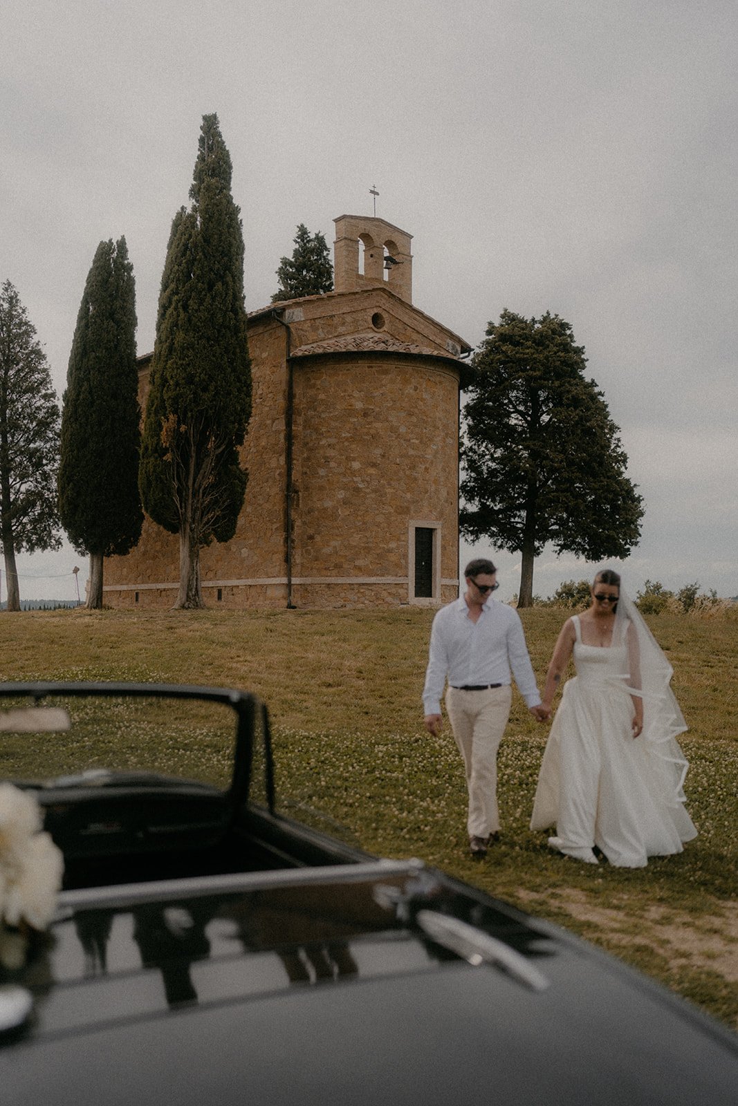A bride and groom walking hand in hand through a grassy area with a small stone church and tall trees in the background, as viewed from a car window.