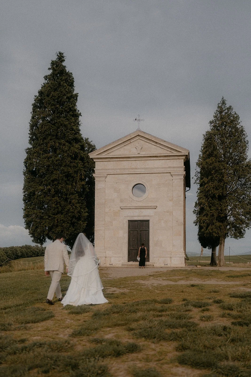 A couple dressed in wedding attire walking towards a small church with tall trees on either side, under a cloudy sky.