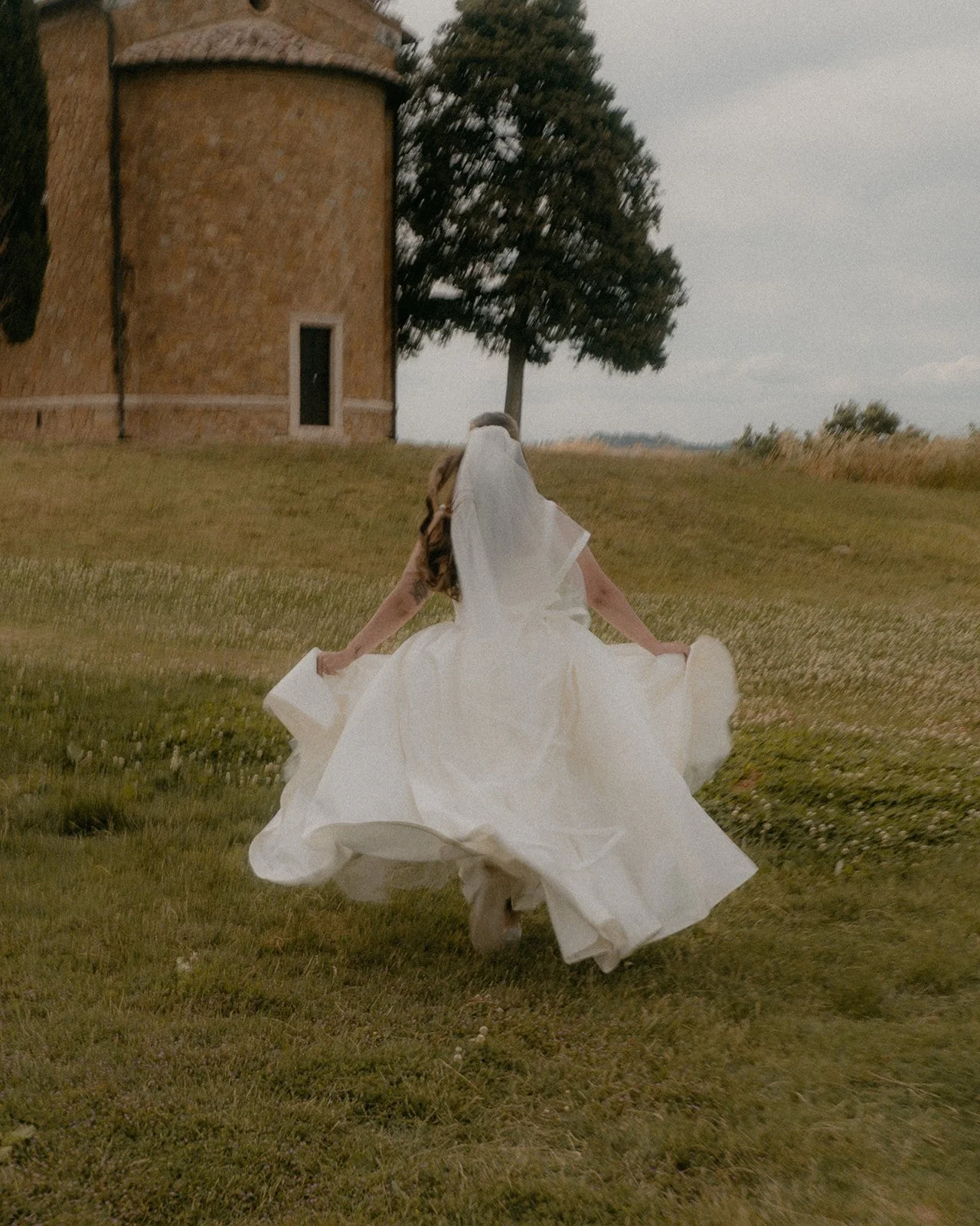 A bride in a white wedding dress and veil running across a grassy field near a stone building and a large tree.
