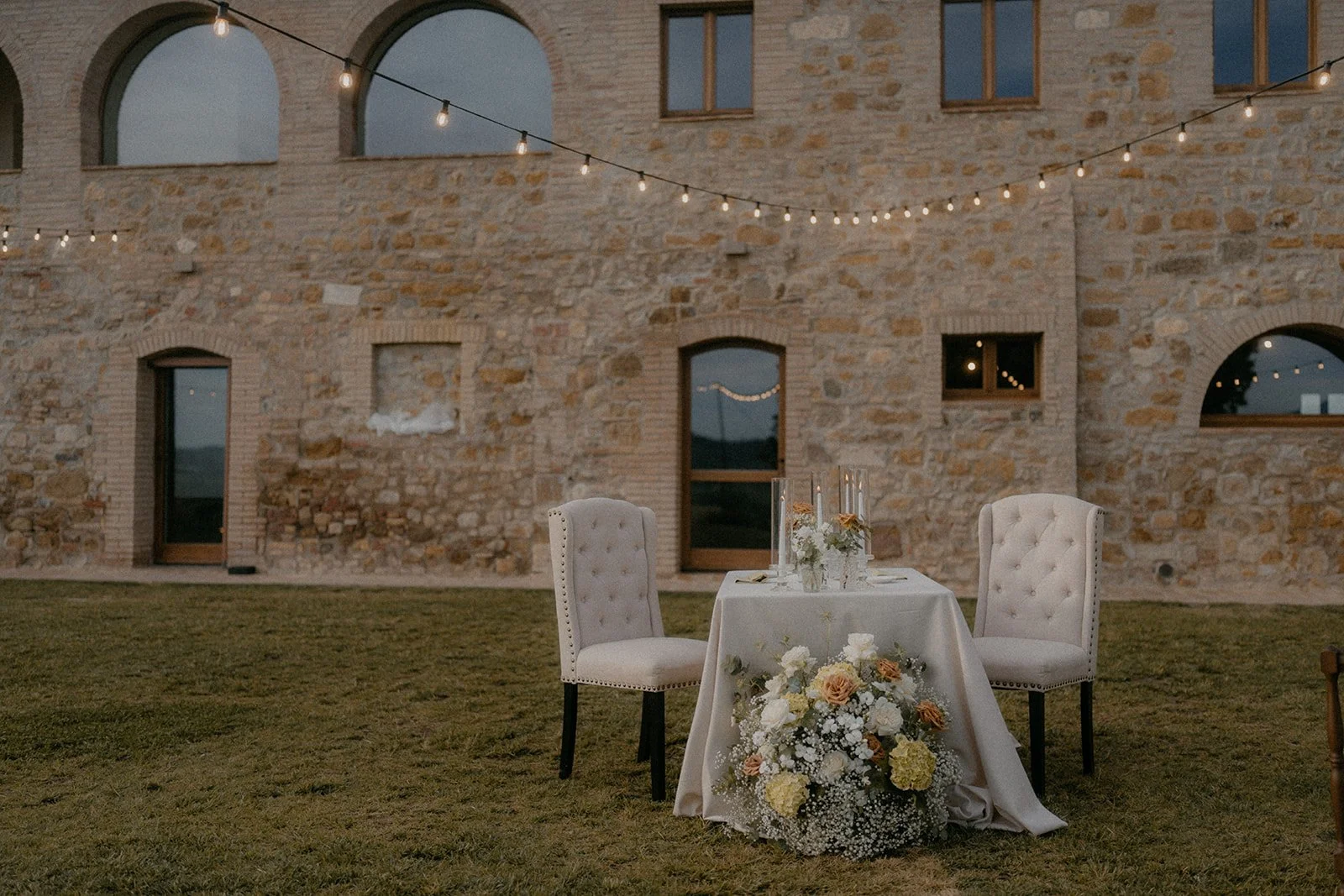 A table set for two with white chairs, floral arrangements, and candles, outside on grass in front of a stone building with string lights overhead during dusk.