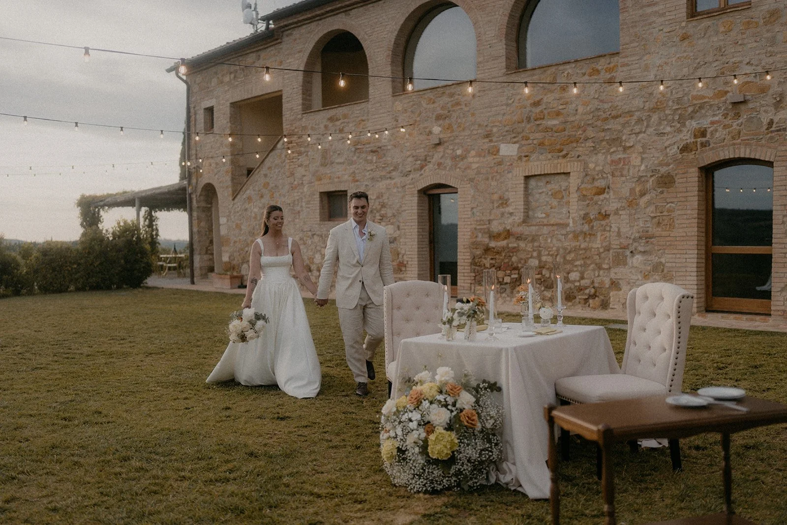 A newlywed couple is walking hand in hand outside a rustic barn-style venue at sunset, with string lights overhead, a table with white tablecloth, floral arrangements, and chairs in the foreground.