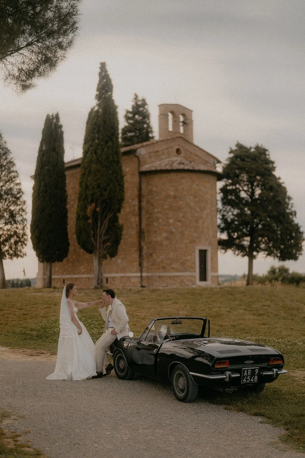A bride and groom are outside near a vintage black convertible car, with a rustic stone building and tall cypress trees in the background during sunset.
