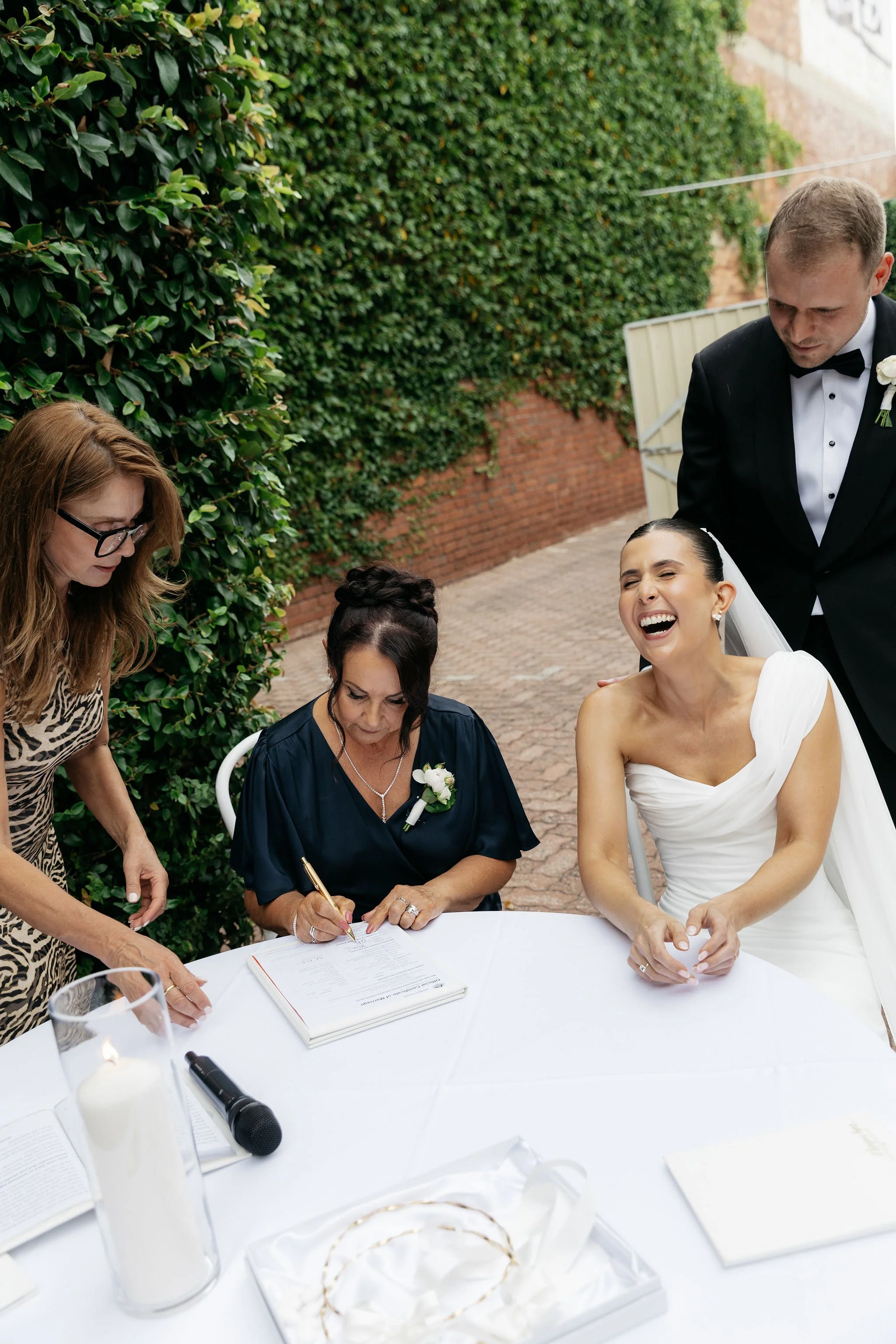 Irene Palamaras showing witness where to sign and bride laughing at signing table with groom standing behind her
