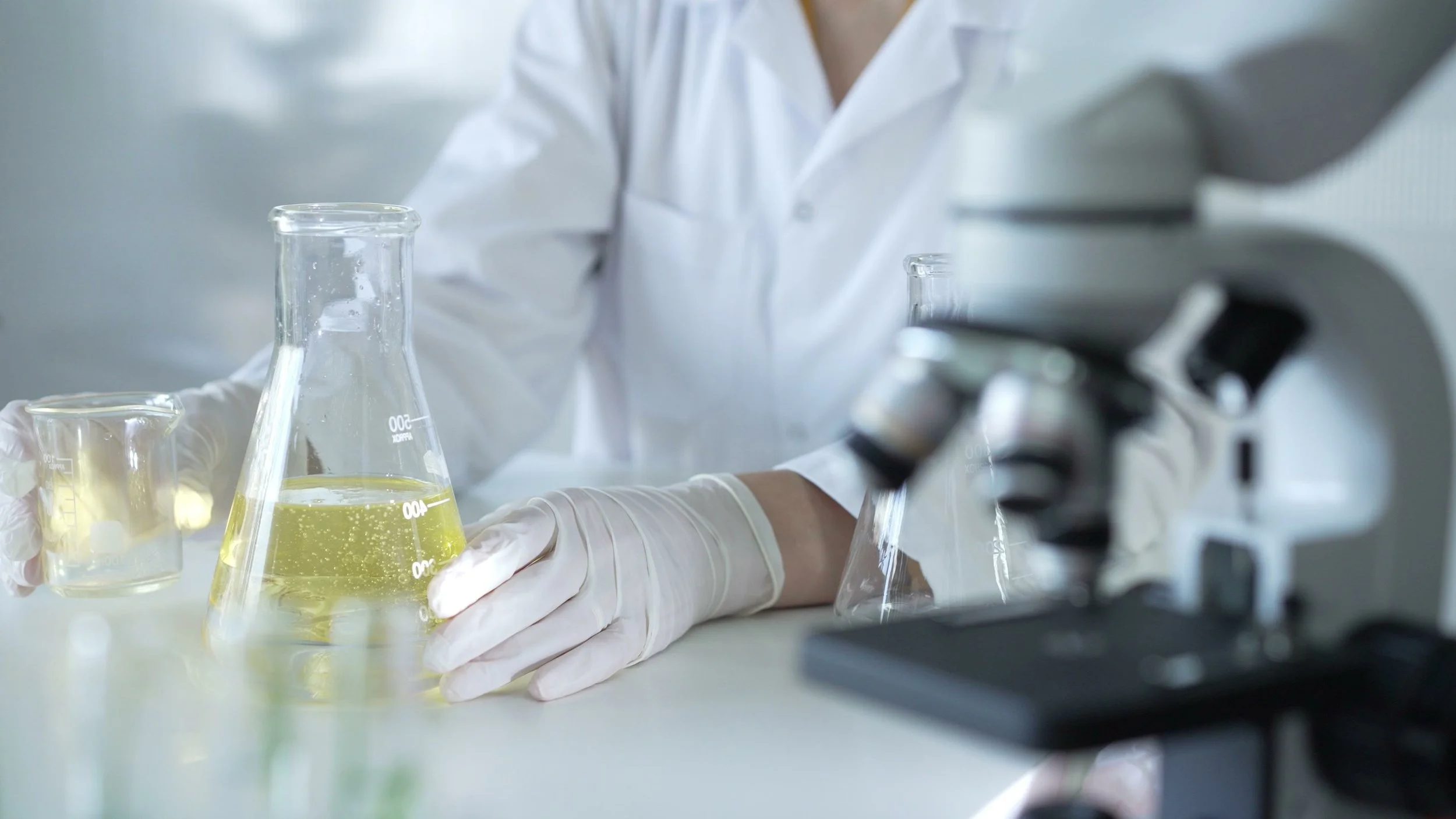 Scientist in a laboratory wearing gloves, handling a beaker with a yellow liquid, with laboratory glassware and a microscope around.