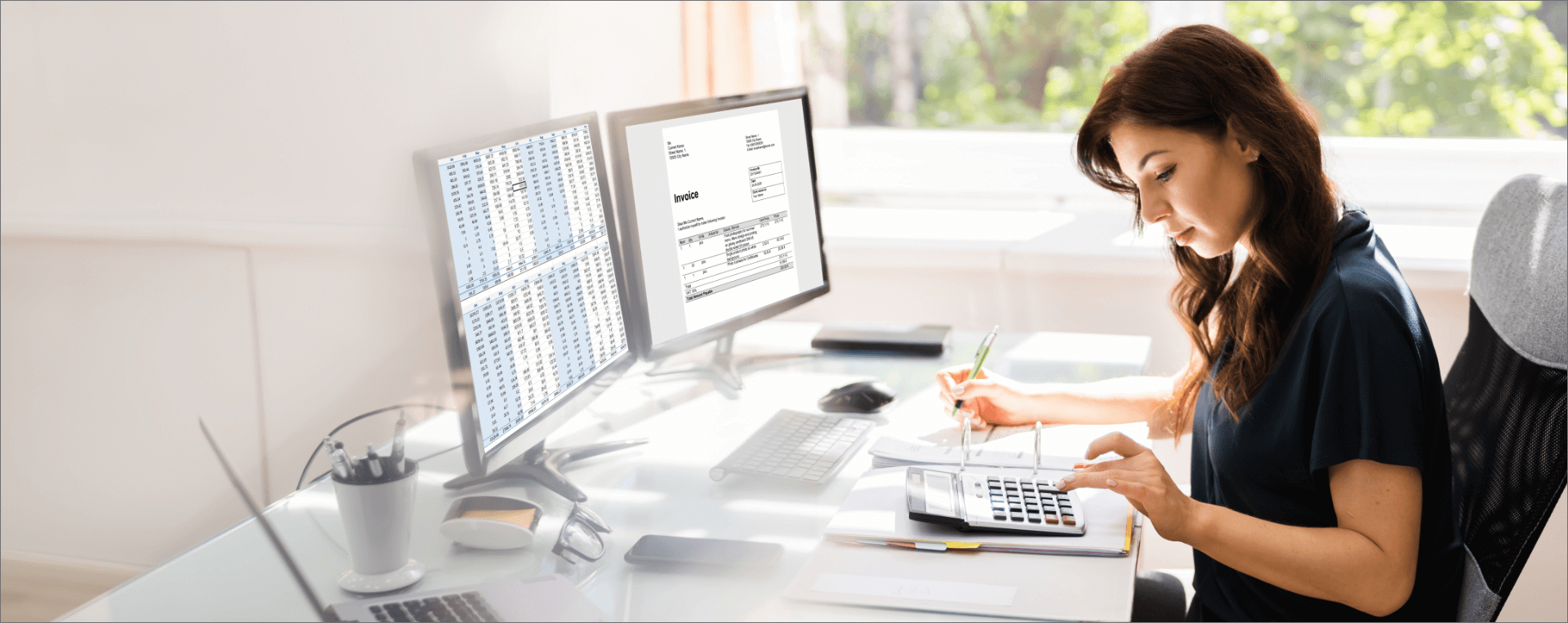 Woman working at a desk with two computer monitors, one displaying spreadsheets and the other a billing invoice, using a calculator, and taking notes with a pen.