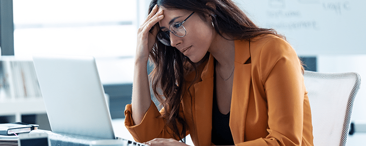 A woman with long brown hair in glasses and an orange blazer sitting at a desk working on a laptop with a white board in the background.