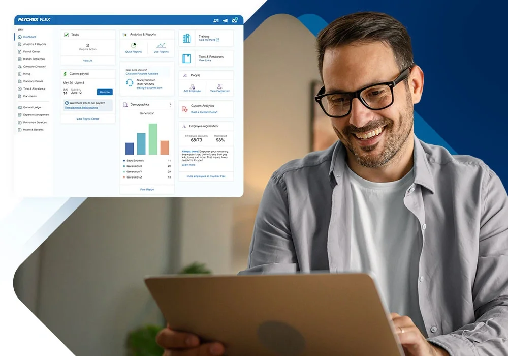 A man with glasses and a beard smiling while looking at a tablet. In the background, there is a computer screen displaying payroll and analytics software.