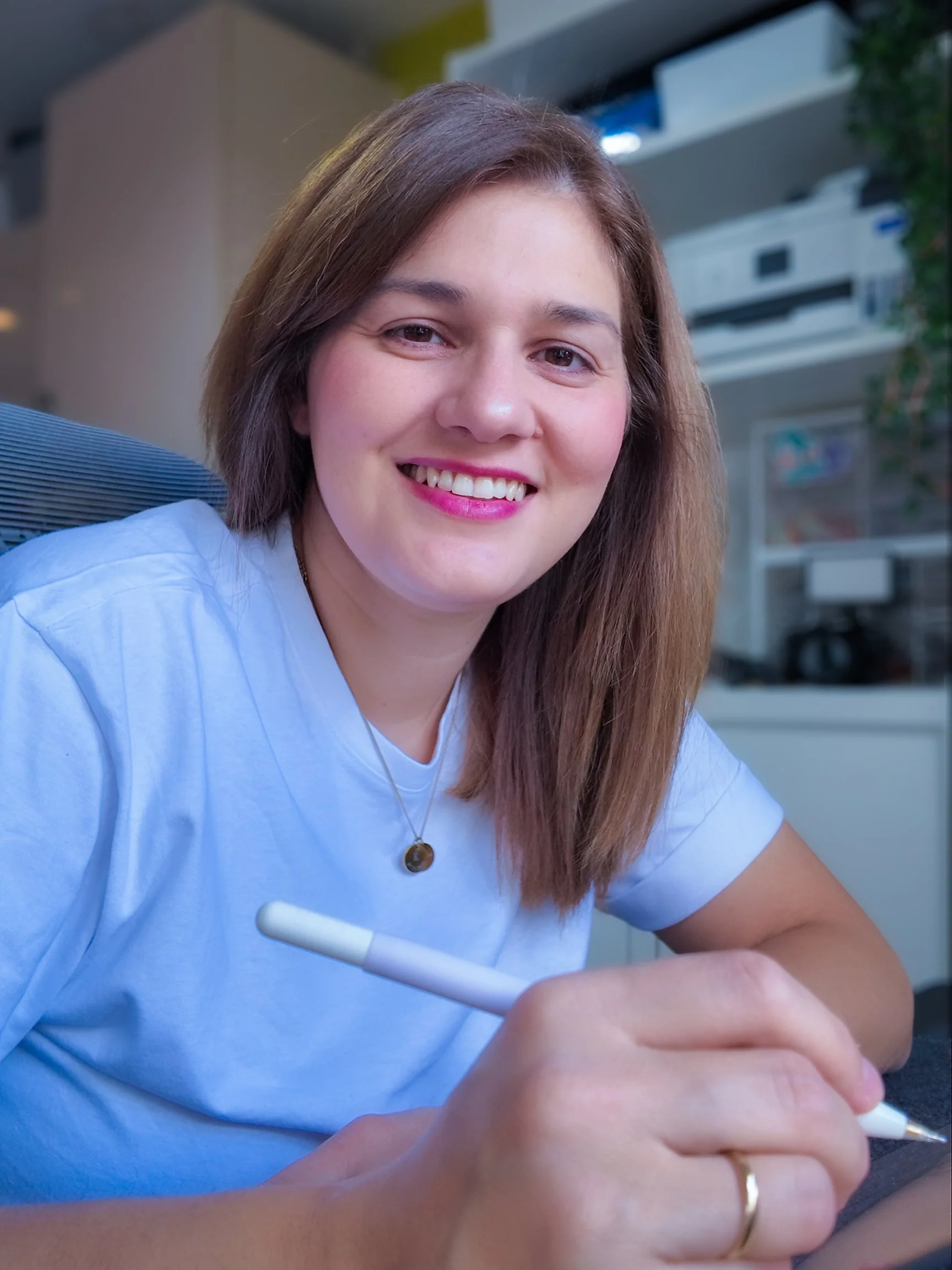 A woman with shoulder-length brown hair smiles while writing with a white pen, wearing a white shirt and a necklace, in a modern indoor setting.