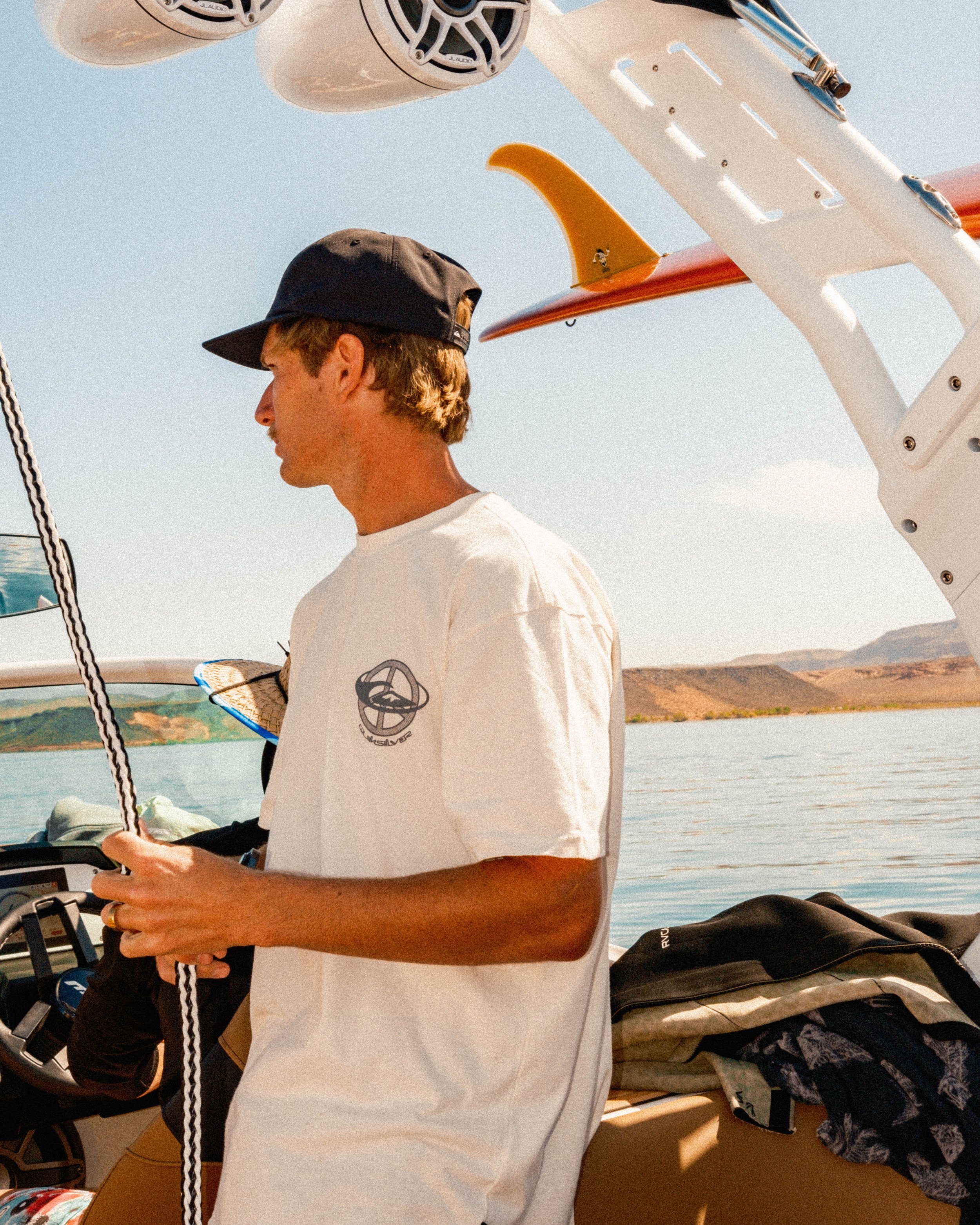 Young man on a boat holding a rope, wearing a white T-shirt and a black cap, with a water landscape and mountains in the background.