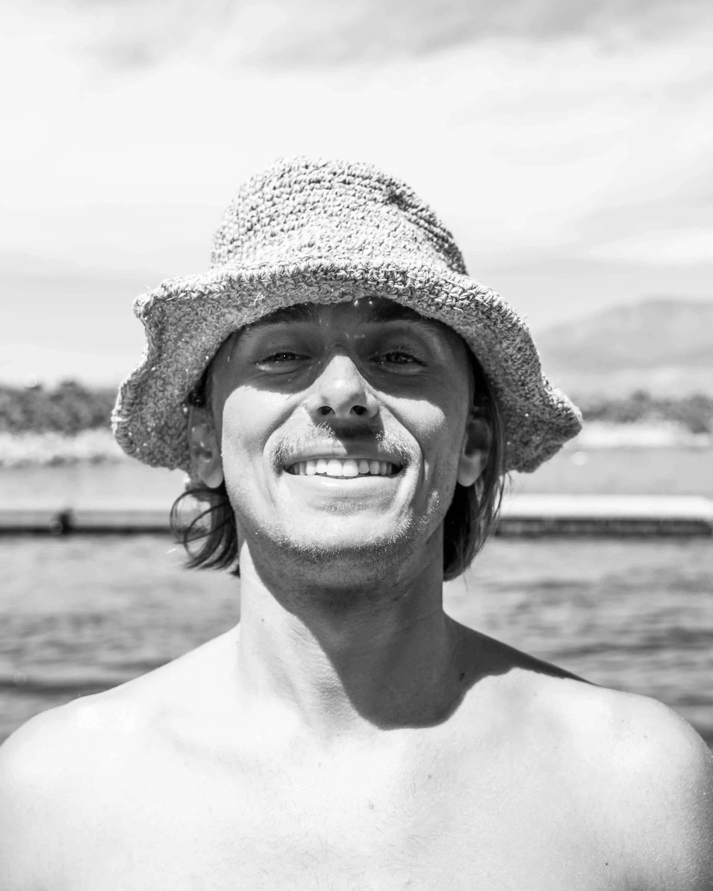 A shirtless man wearing a straw hat outdoors on a sunny day, smiling at the camera, with water and landscape in the background.