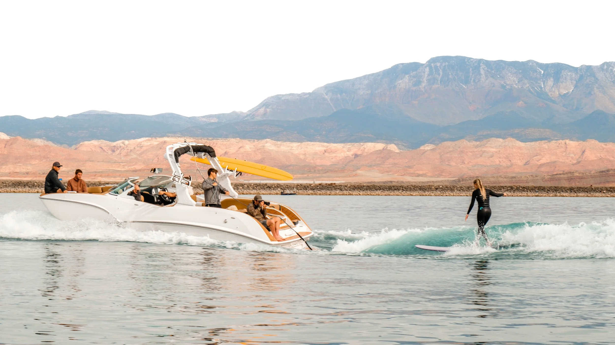 A woman surfing on a wakeboard behind a speeding boat on a lake, with mountains in the background.