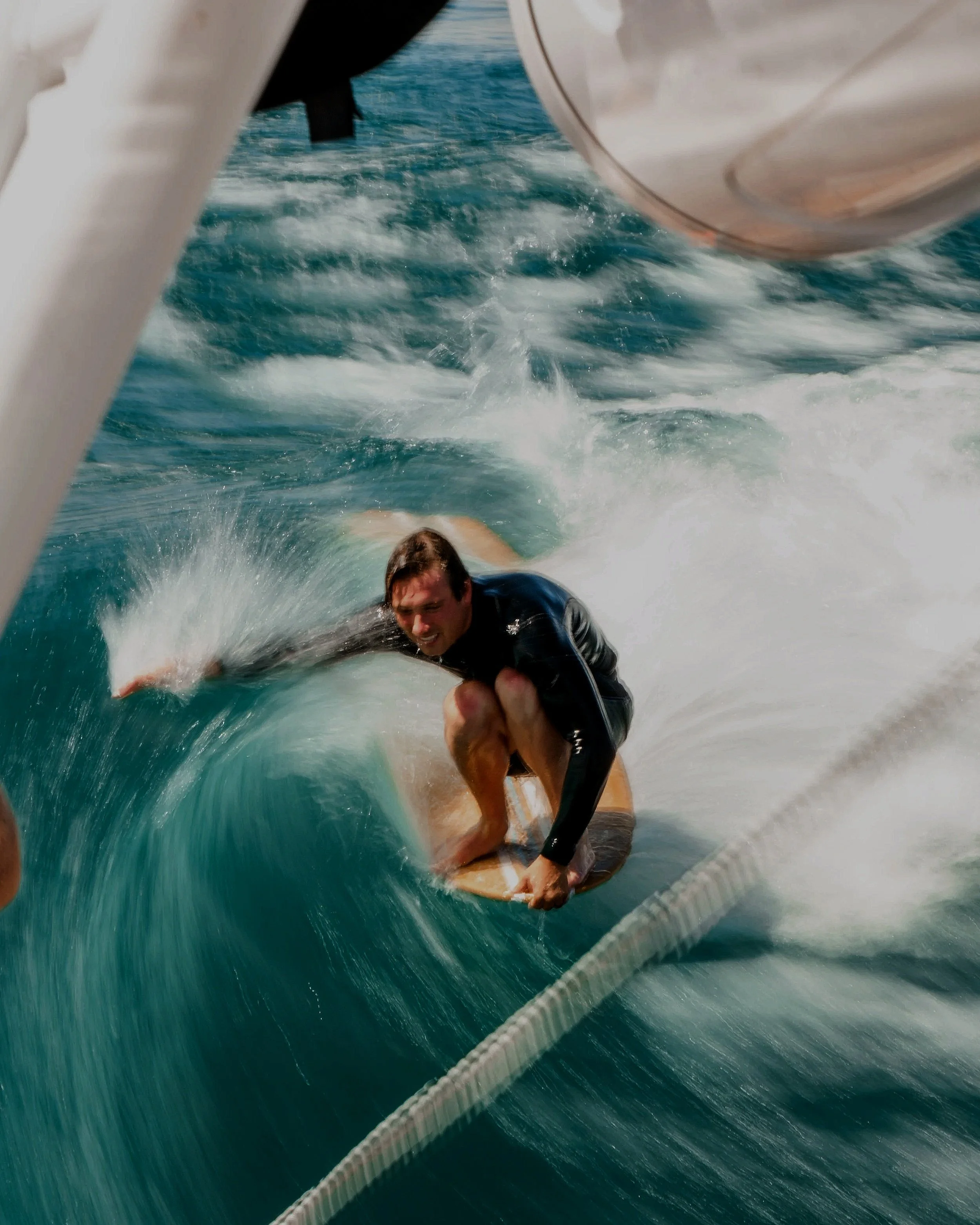 Man surfing on a wave while wearing a black wetsuit, captured from a boat in the water.