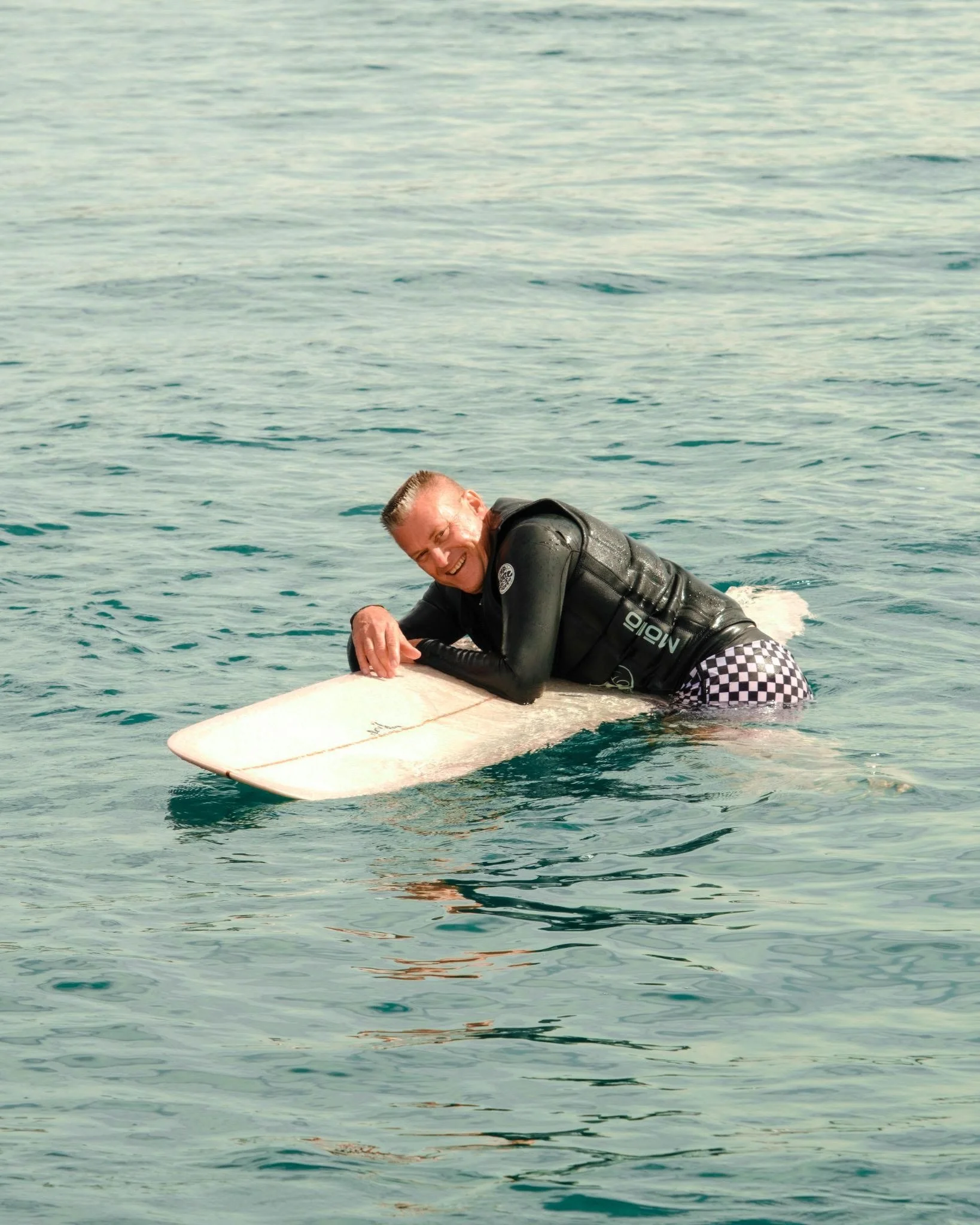 A man smiling while lying on a surfboard in the water, wearing a black wetsuit with checkered shorts.
