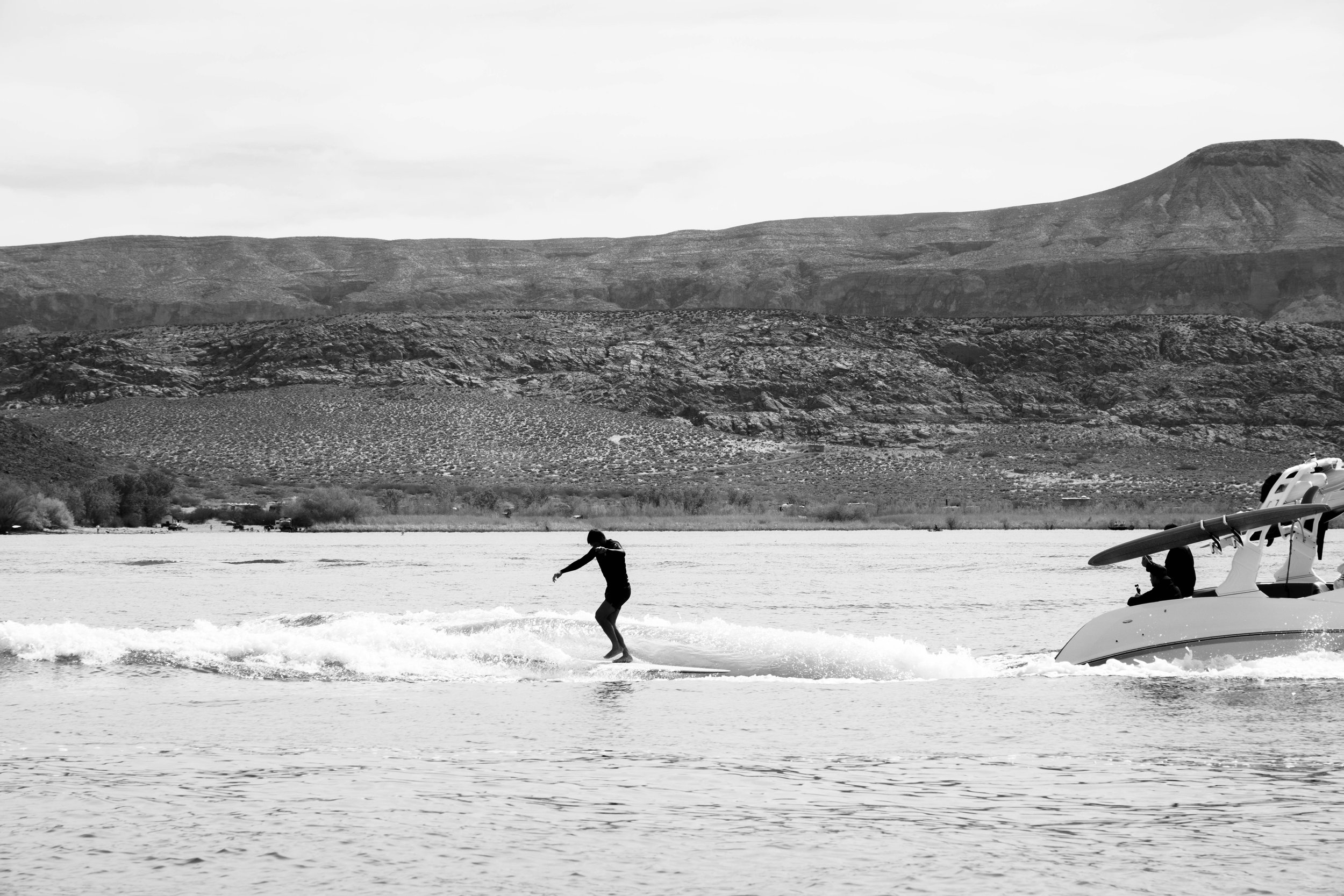 A person surfing on a lake with a boat nearby, and mountains in the background.