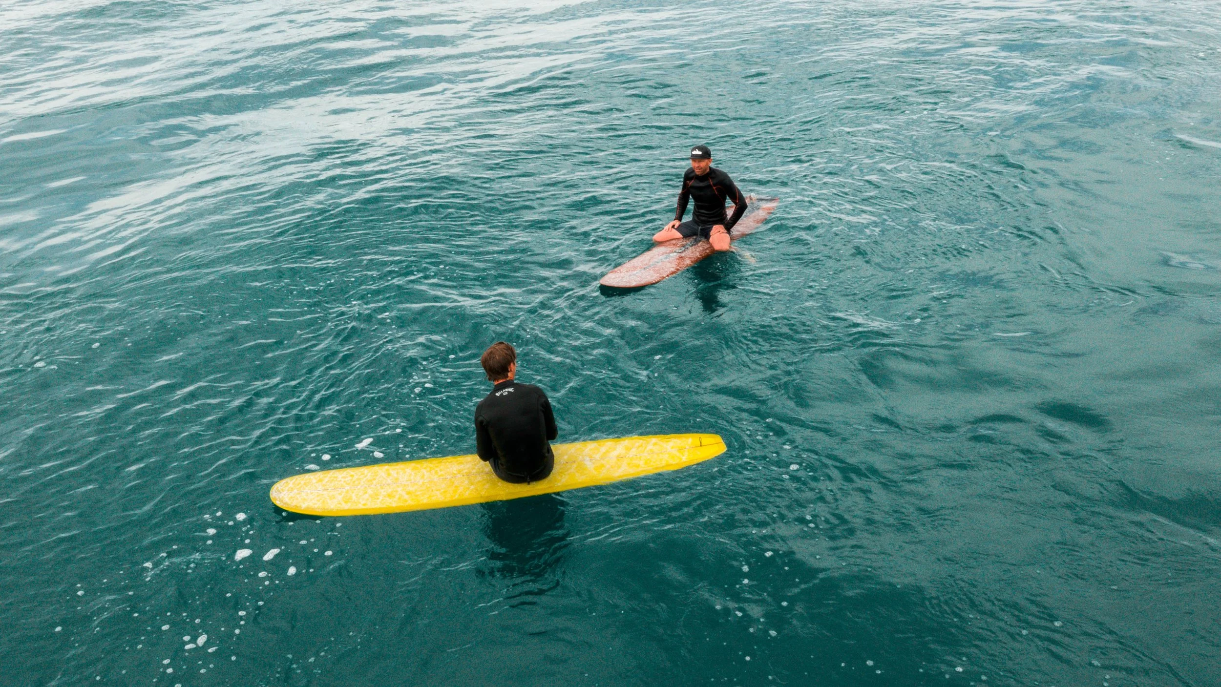 Two surfers in wetsuits on surfboards in the ocean, one sitting on a yellow surfboard and the other kneeling on a brown surfboard, engaging in surfing conversation.