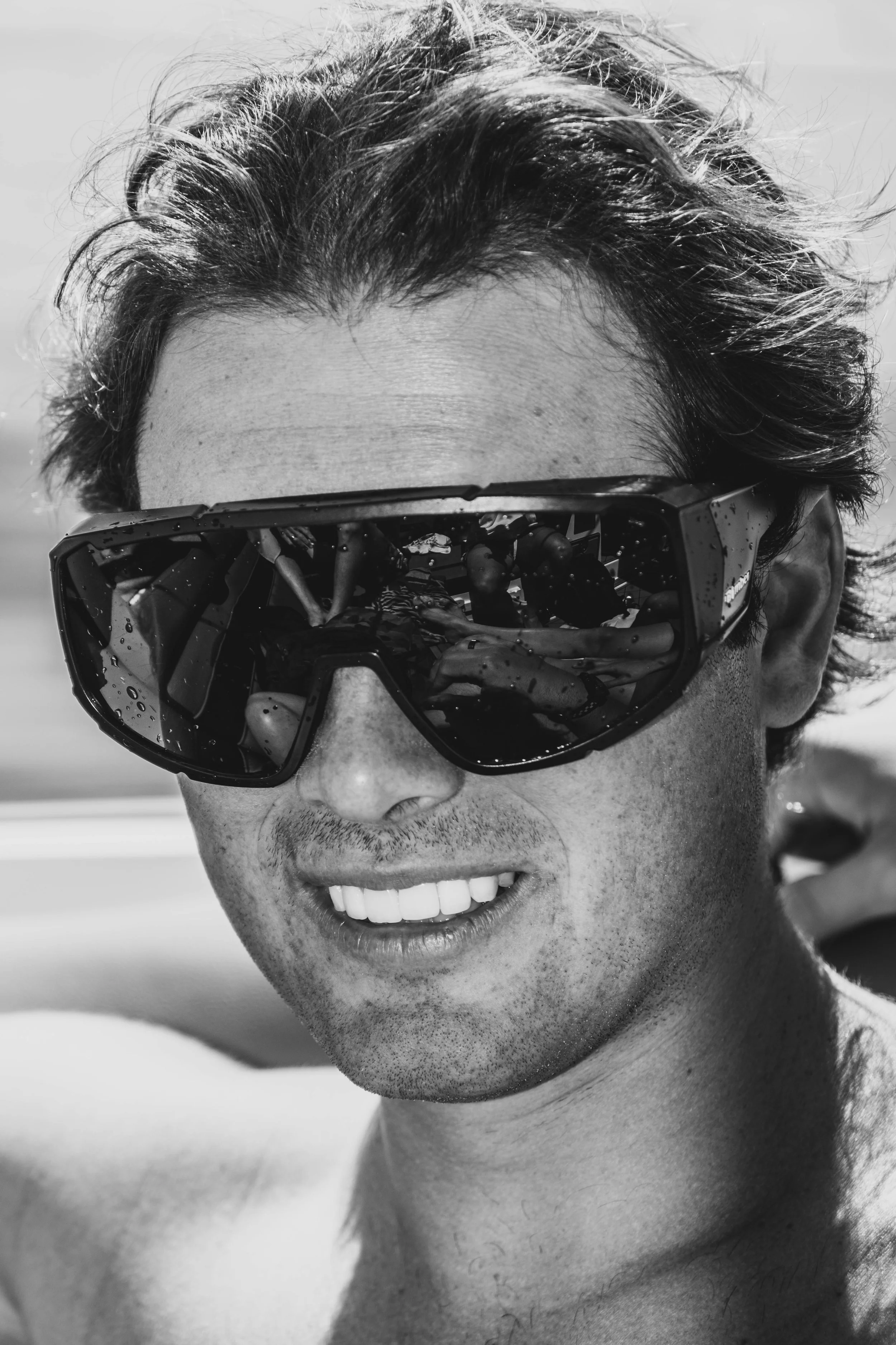 A man smiling while wearing sports sunglasses, with his hair slightly messy and a cloudy sky in the background.