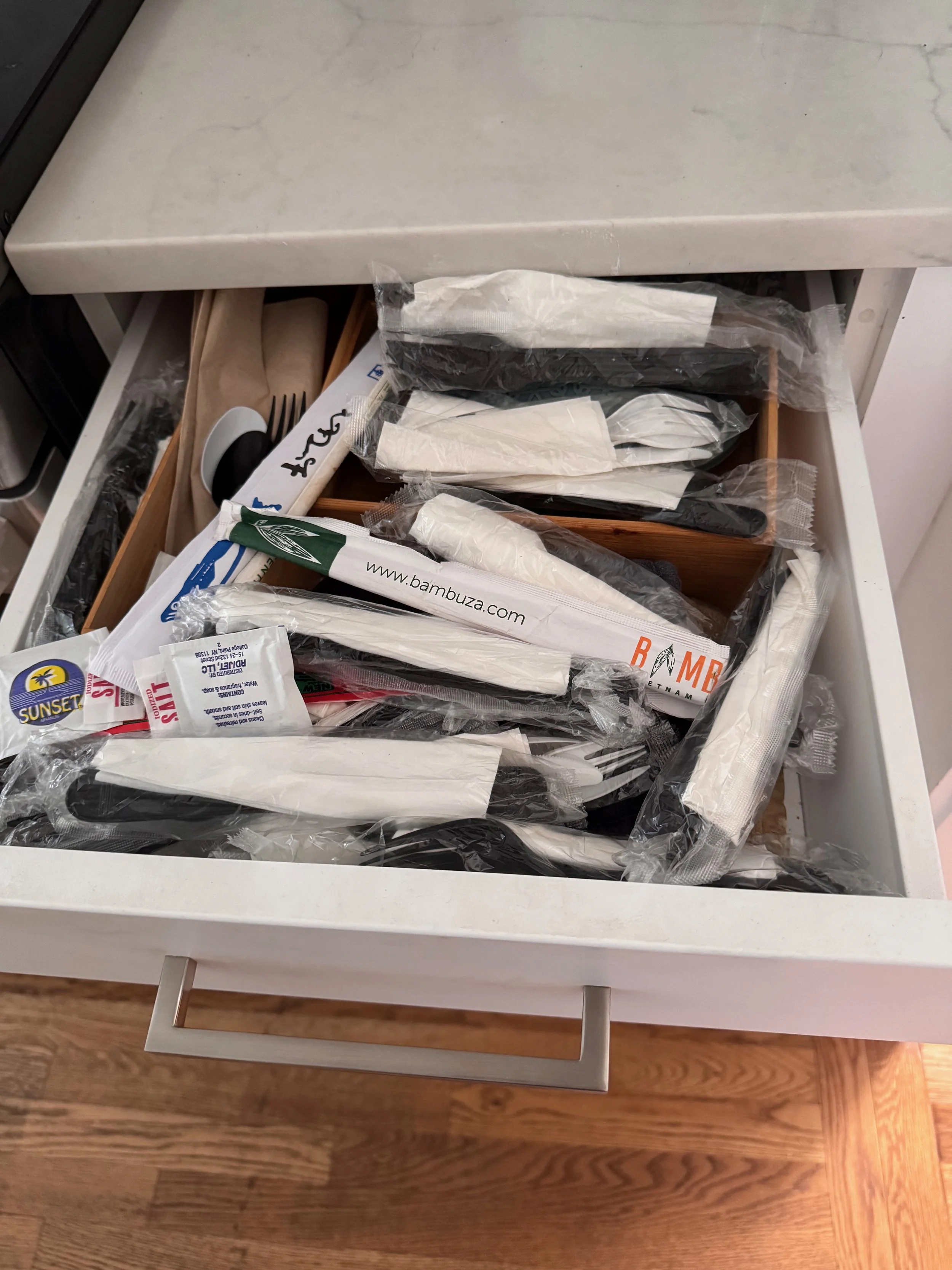 Image shows a kitchen drawer partially open and filled with plastic packets of wrapped plastic forks, spoons, knives, as well as chopsticks and napkins and handwash packets.