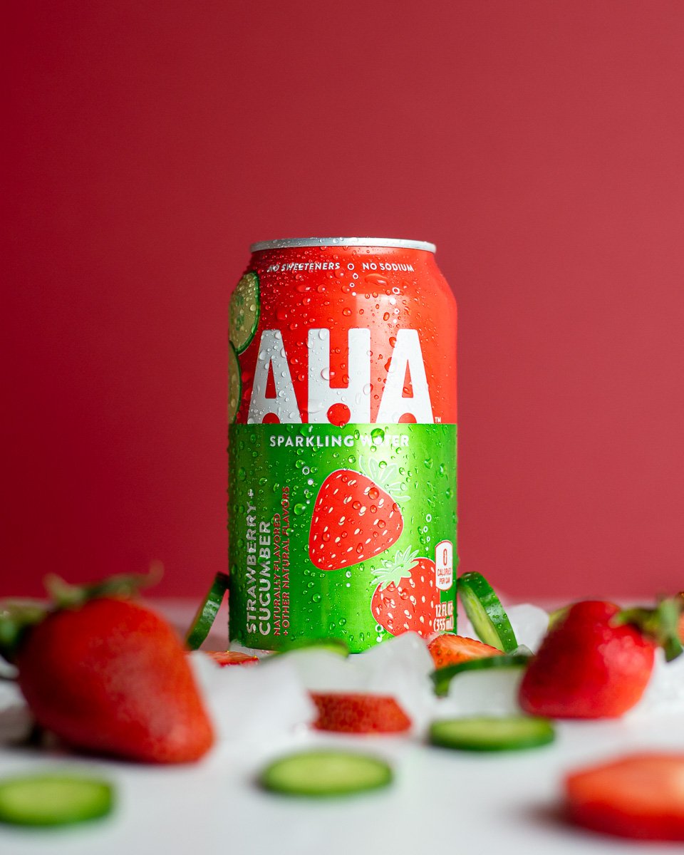 A can of Aha sparkling strawberry cucumber flavored water, surrounded by strawberries and cucumber slices, with a pink background.