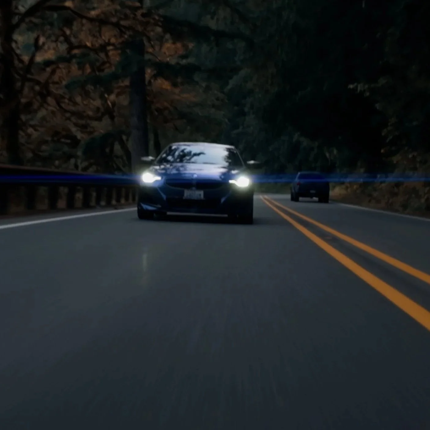 A black sports car with its headlights on driving down a two-lane road surrounded by trees at dusk or dawn.