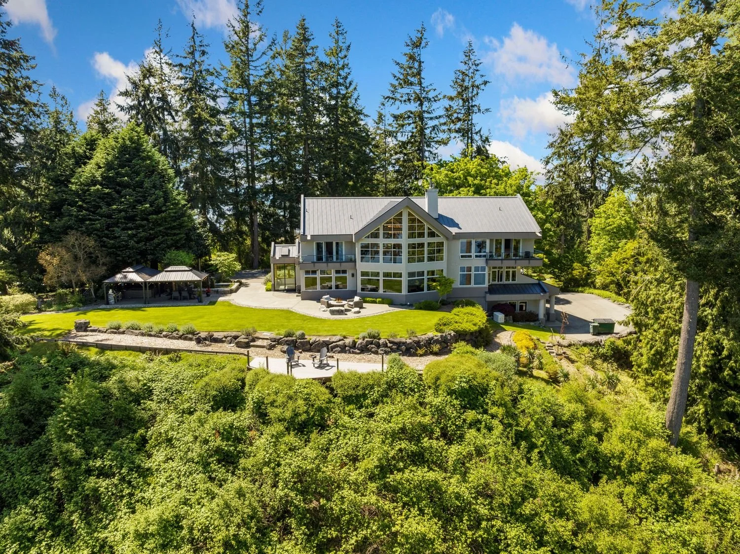 Large modern house with multiple stories, large windows, and a gray roof surrounded by lush greenery and tall trees on a sunny day.