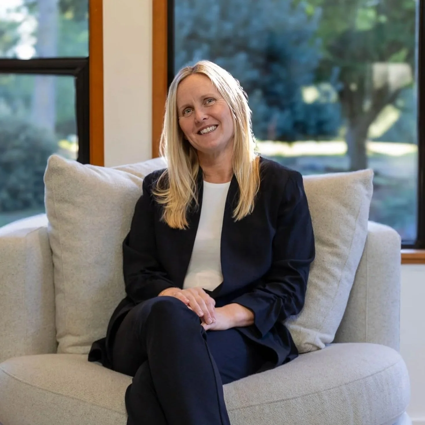 A woman with long blonde hair, wearing a black blazer and white shirt, sitting on a beige couch in front of large windows revealing a green outdoor landscape.